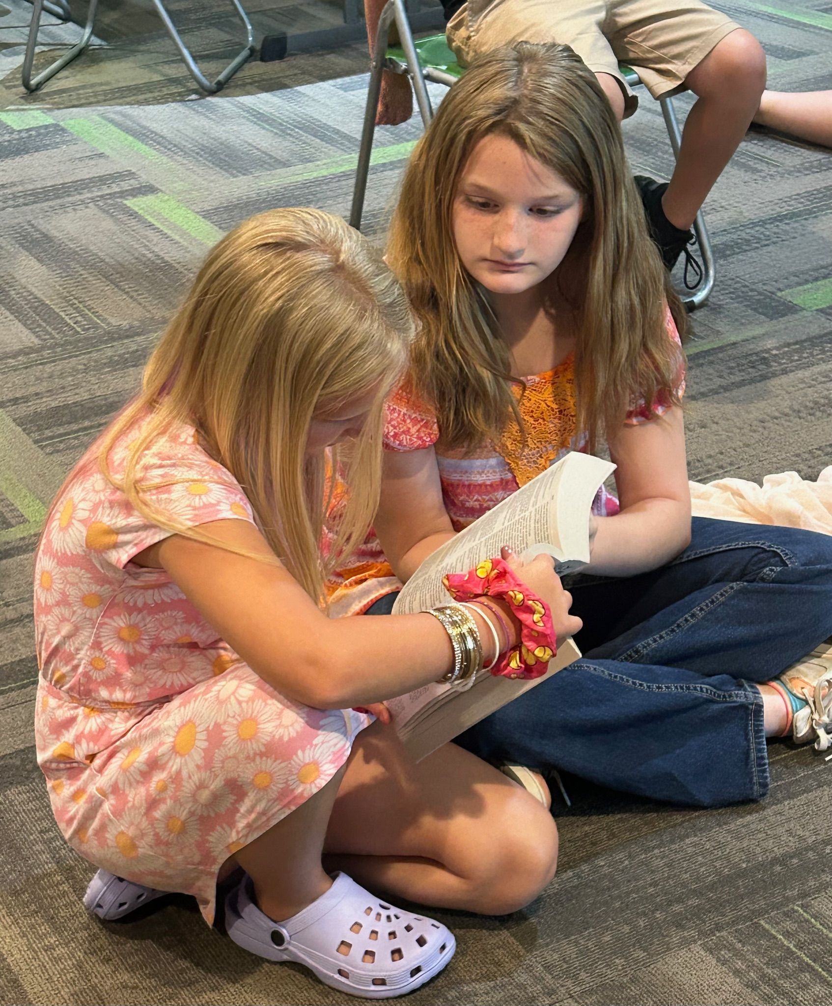 Two children sit on the floor, reading a book together in a carpeted room.
