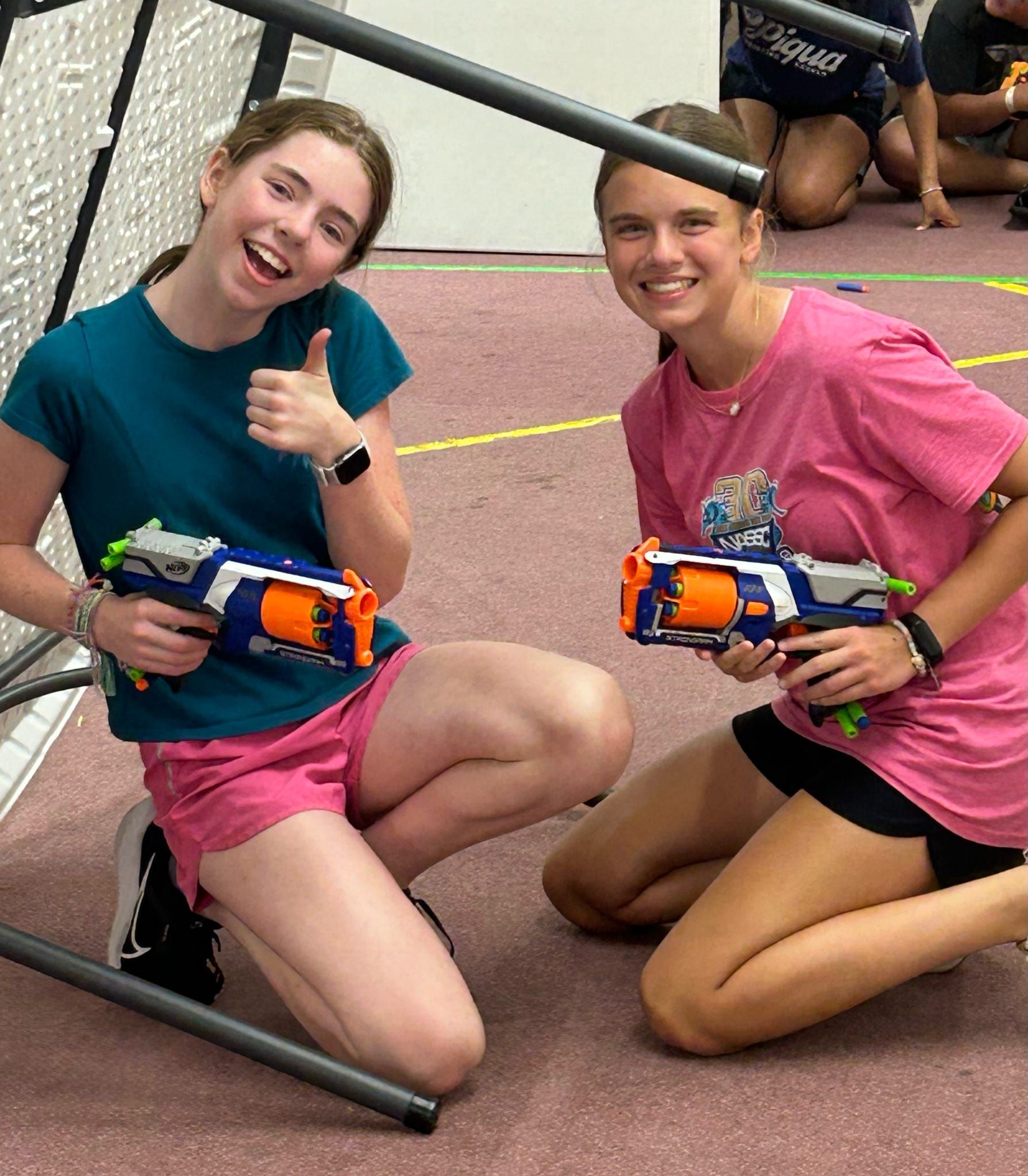 Two smiling people kneel on a gym floor, holding orange toy blasters and posing playfully.