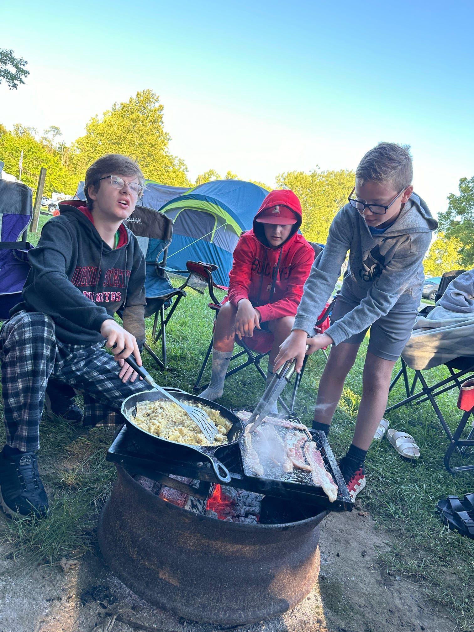 Three campers cooking over a fire pit beside tents in a wooded campsite.