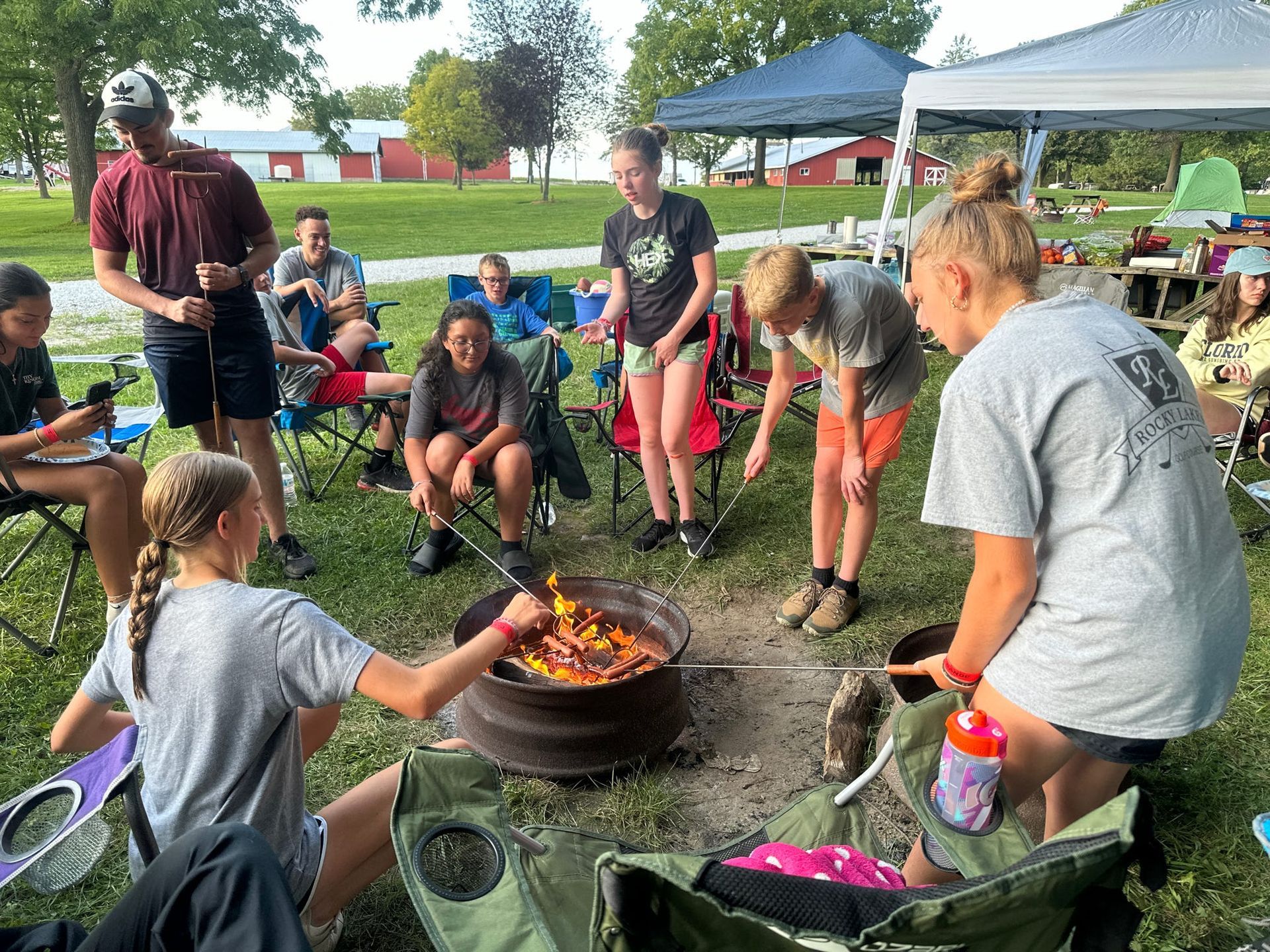 Campers gather around a fire pit, roasting food under a blue canopy in a park.