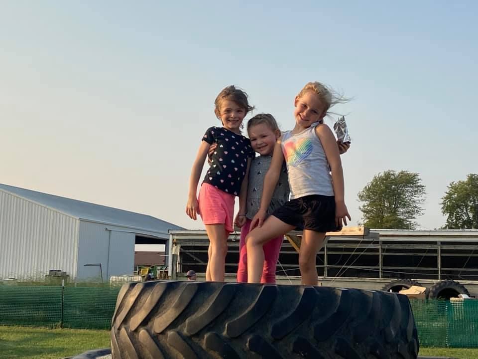 Three children standing on a large tractor tire outdoors near a farm building at sunset