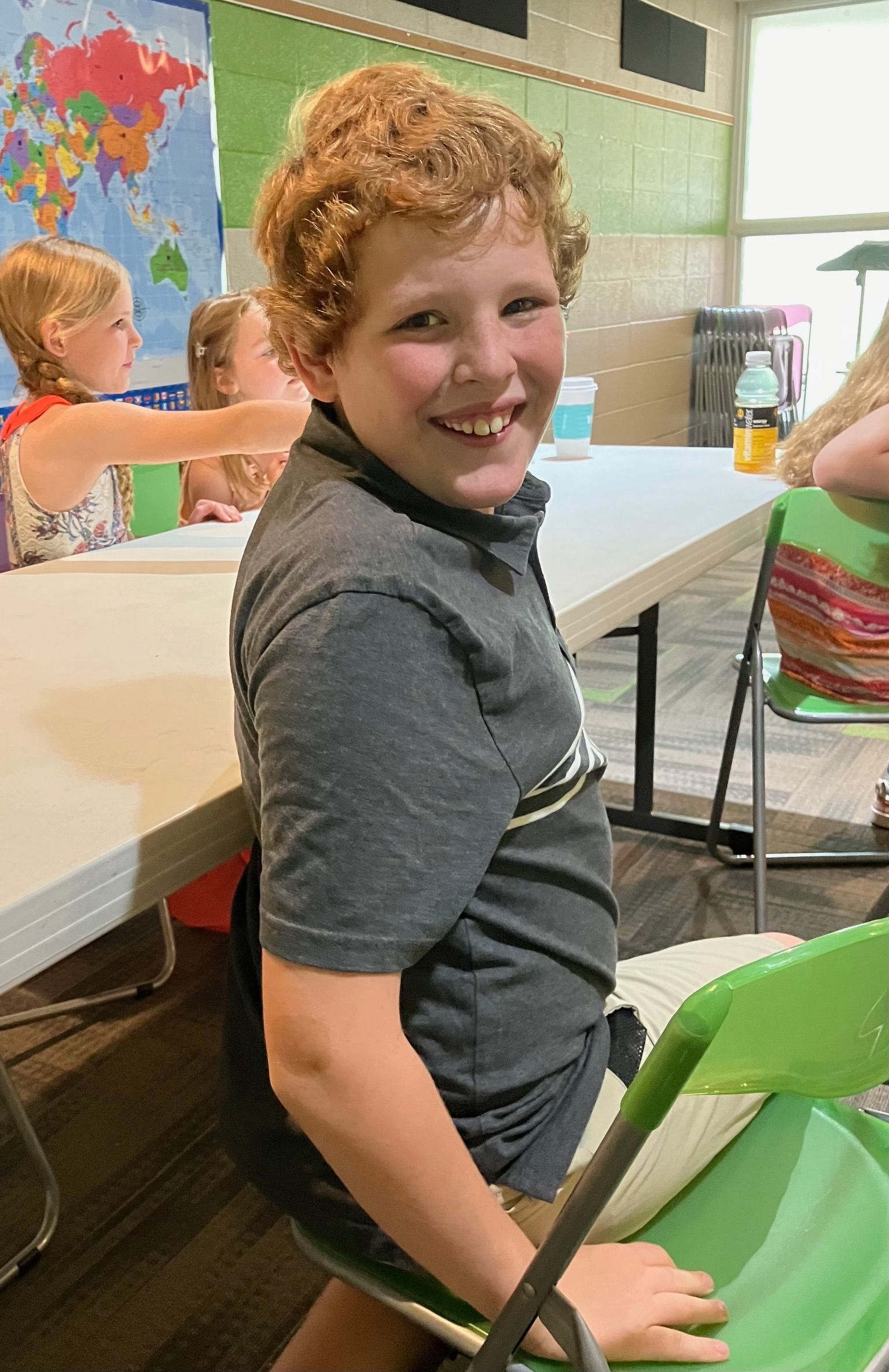 Smiling child in a gray shirt sitting at a table in a classroom, with colorful artwork in the background.