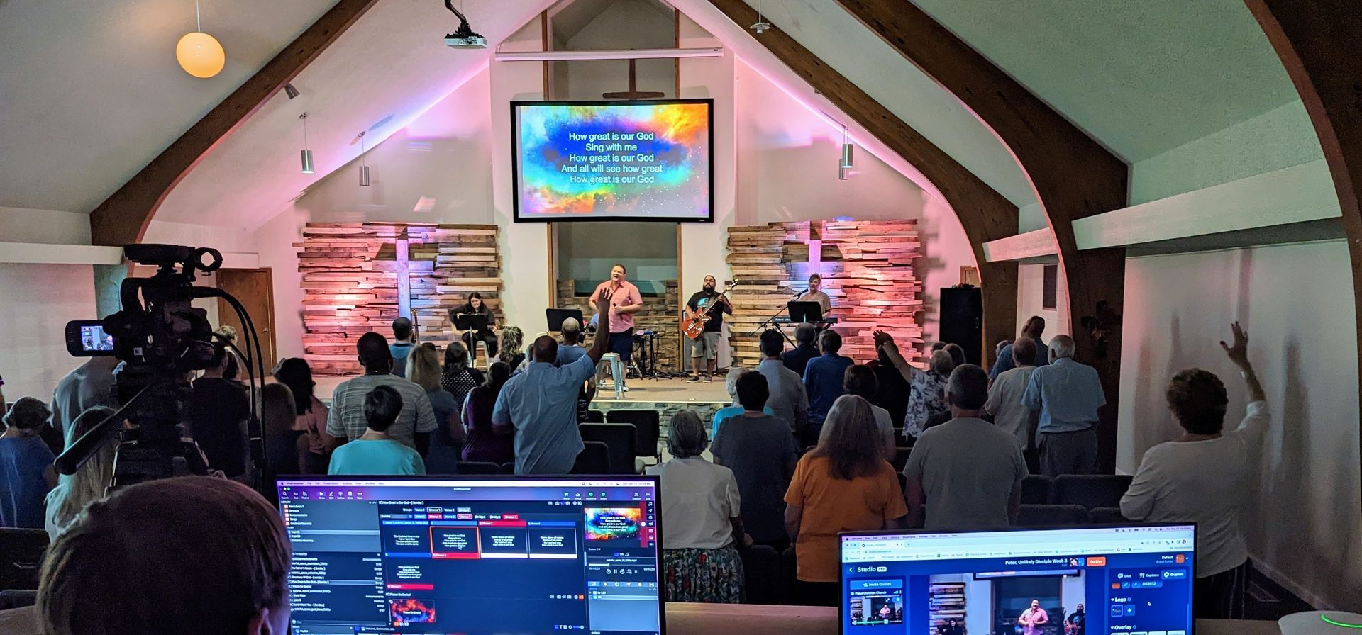 Auditorium worship service with people seated, speakers on stage, and a colorful screen overhead