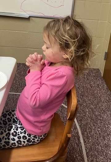 Child in pink top and leopard-print leggings sits on a wooden chair, facing left in a classroom.