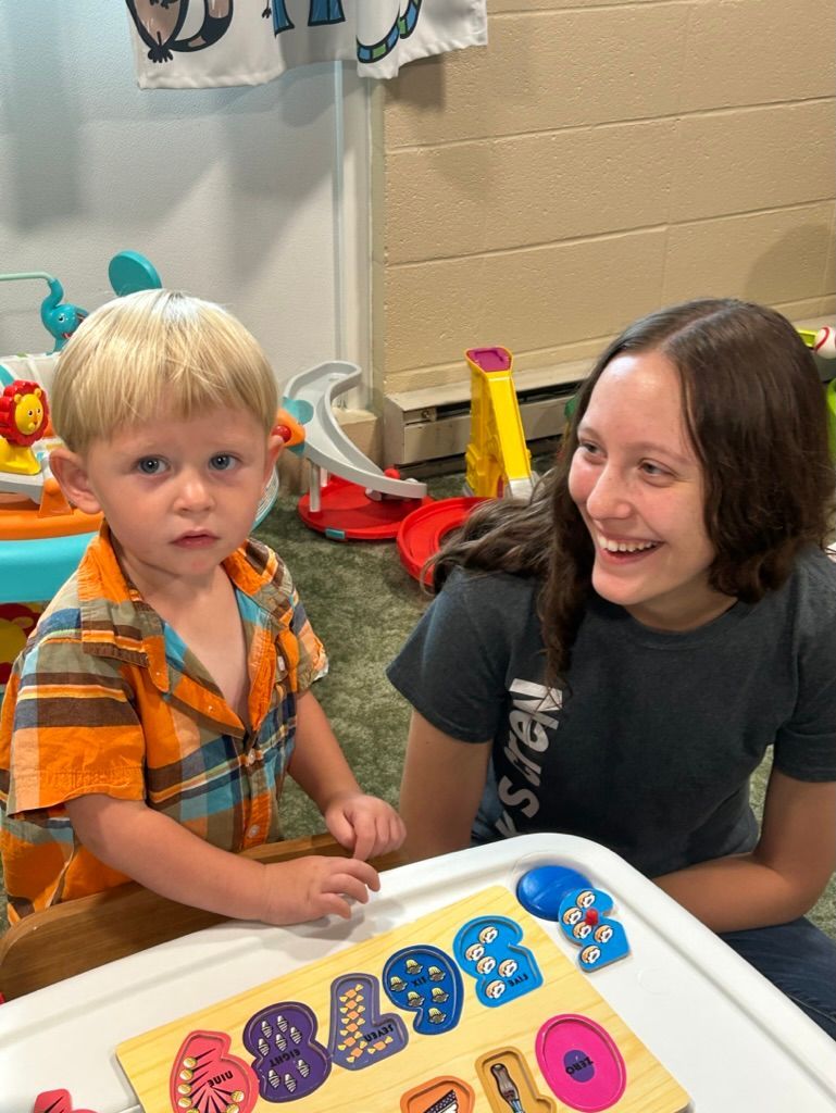 Two children smiling beside a table with colorful letter toys and a beach ball in a classroom