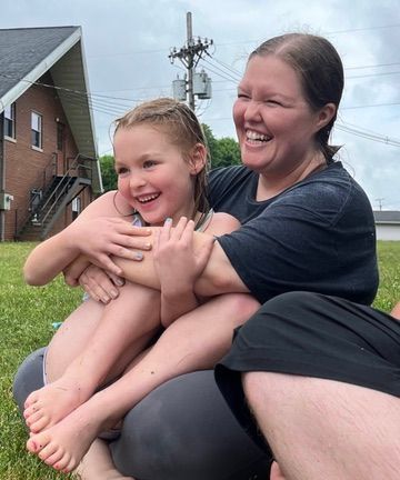 Smiling woman hugging a laughing child outdoors on grass near a building and utility pole
