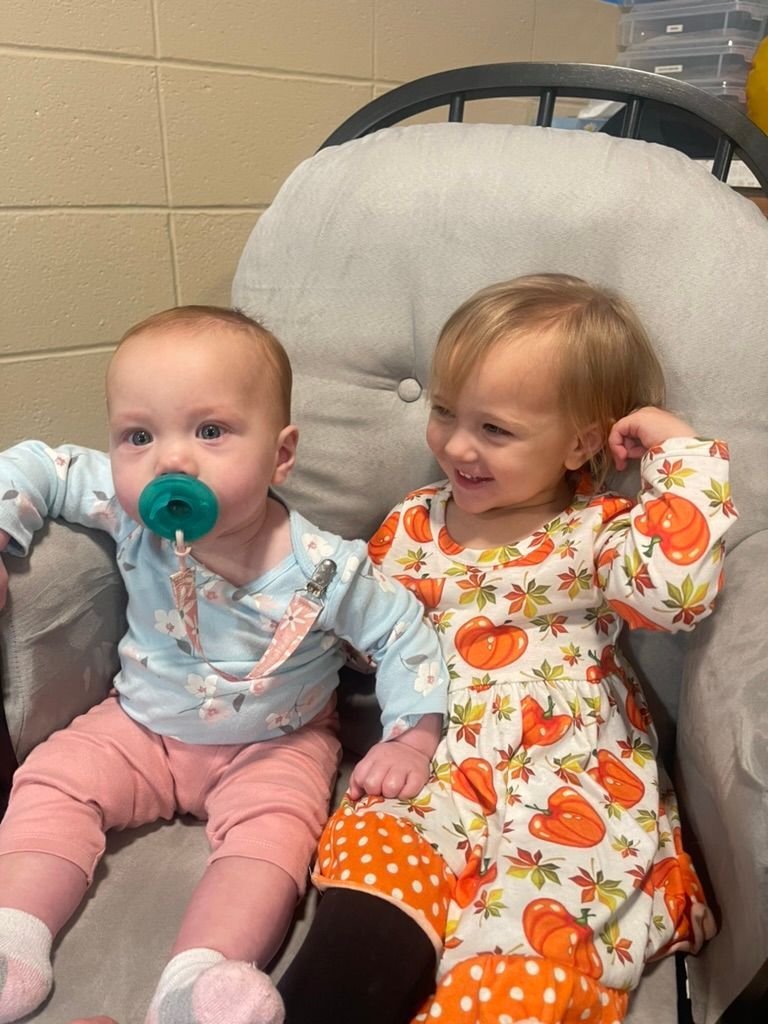 Two babies sitting on a chair, one with a pacifier, the other smiling in a floral outfit