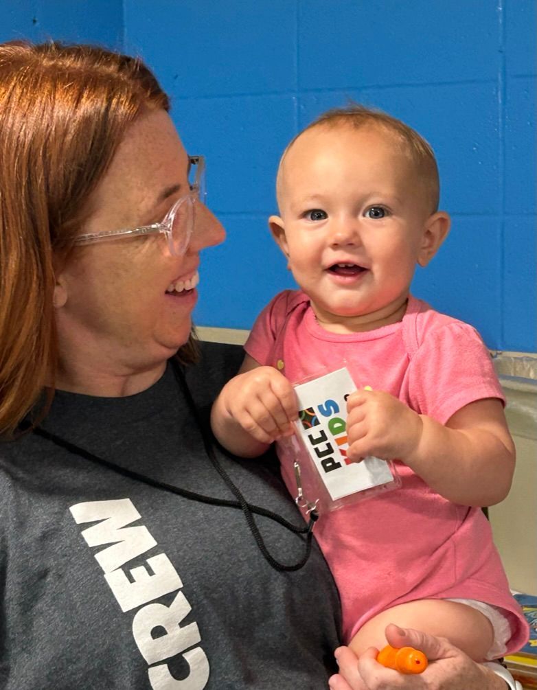 Woman holding smiling baby in pink shirt, indoors against a blue wall.