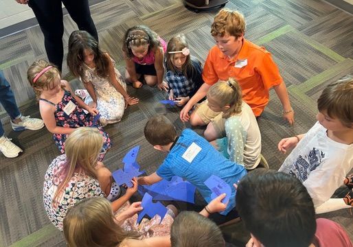 Children gathered in a circle around a person in a bright blue shirt, looking at something on the floor.