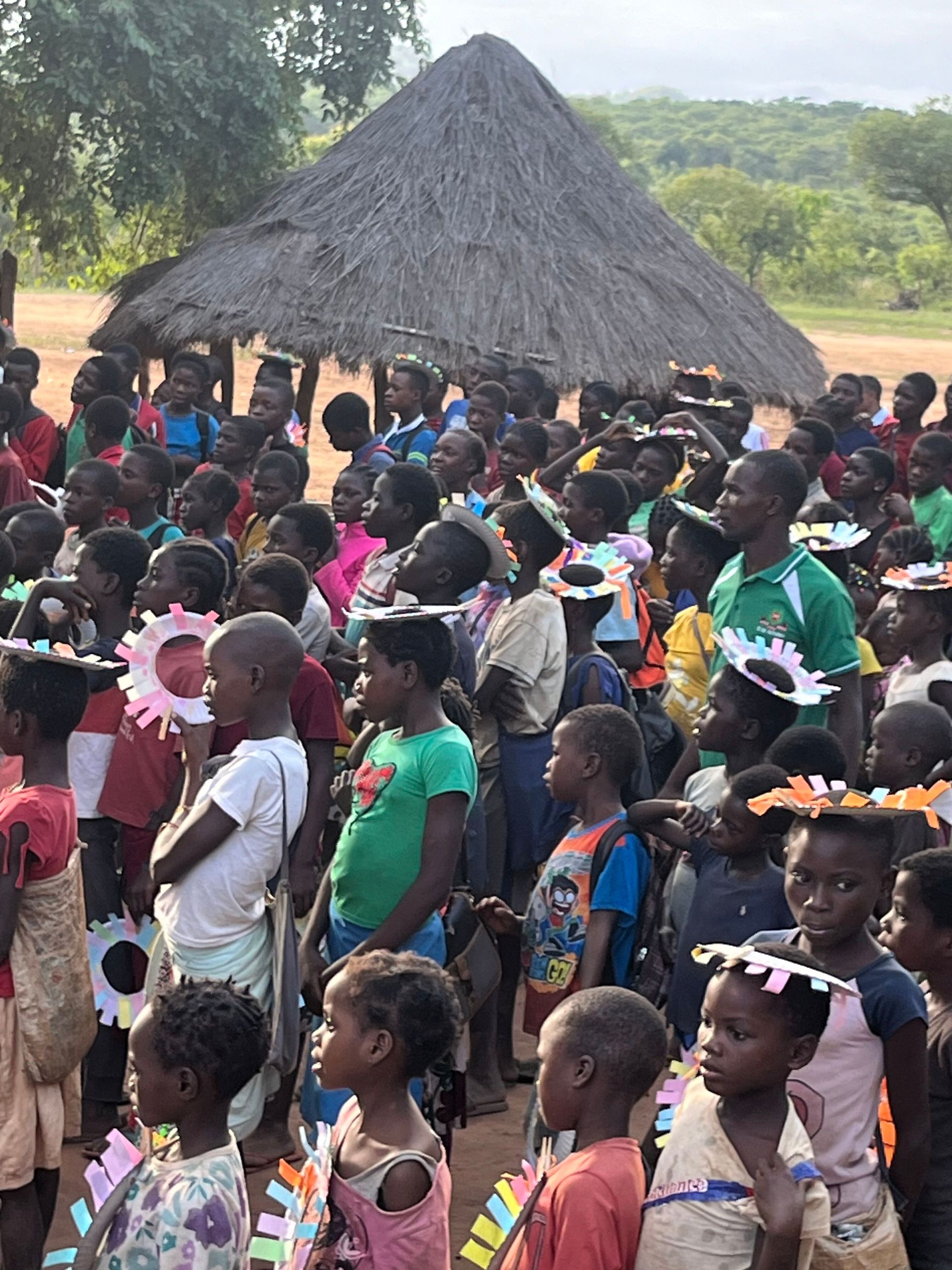 Crowd gathered outdoors near a thatched hut, with people in colorful clothing facing left.