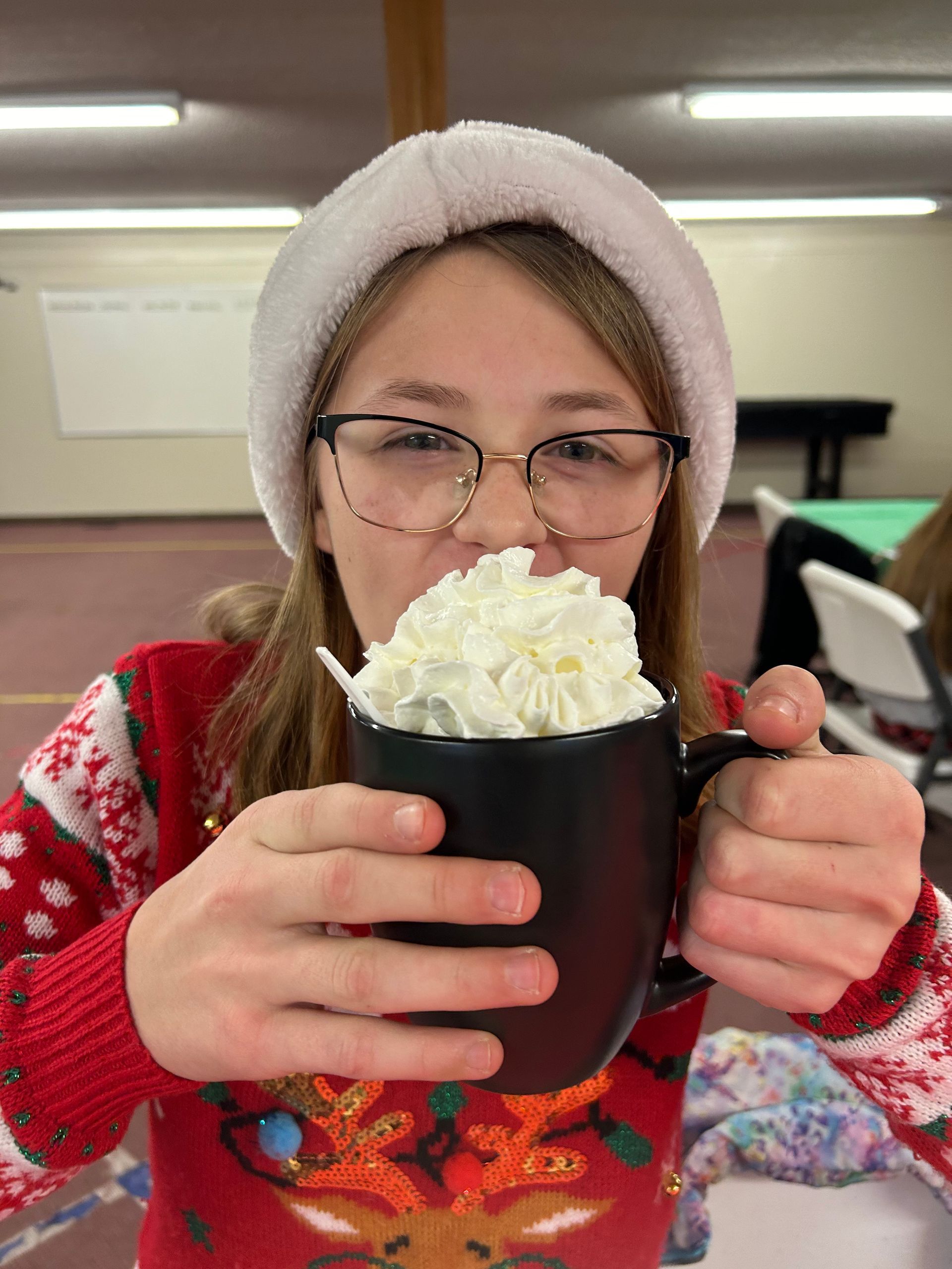 Person in glasses and a knit hat holding a mug topped with whipped cream in a bright indoor room.