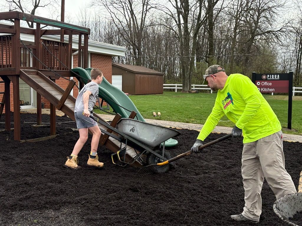 Two people push a wheelbarrow beside a playground on wet black mulch.