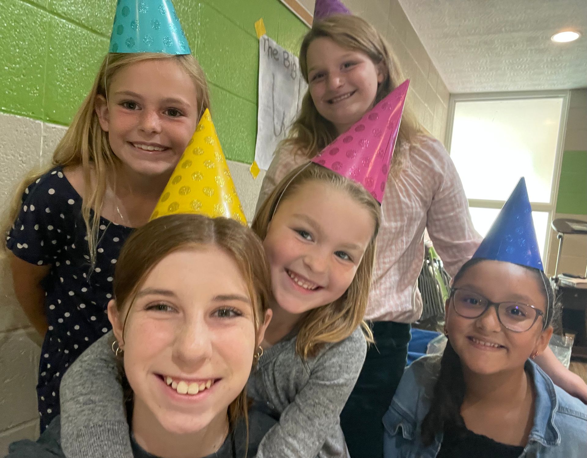 Five smiling children wearing colorful party hats pose in a hallway.