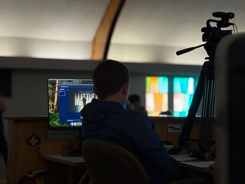 Person seated at a desk, viewing a computer screen in a dim studio with a camera on a tripod.