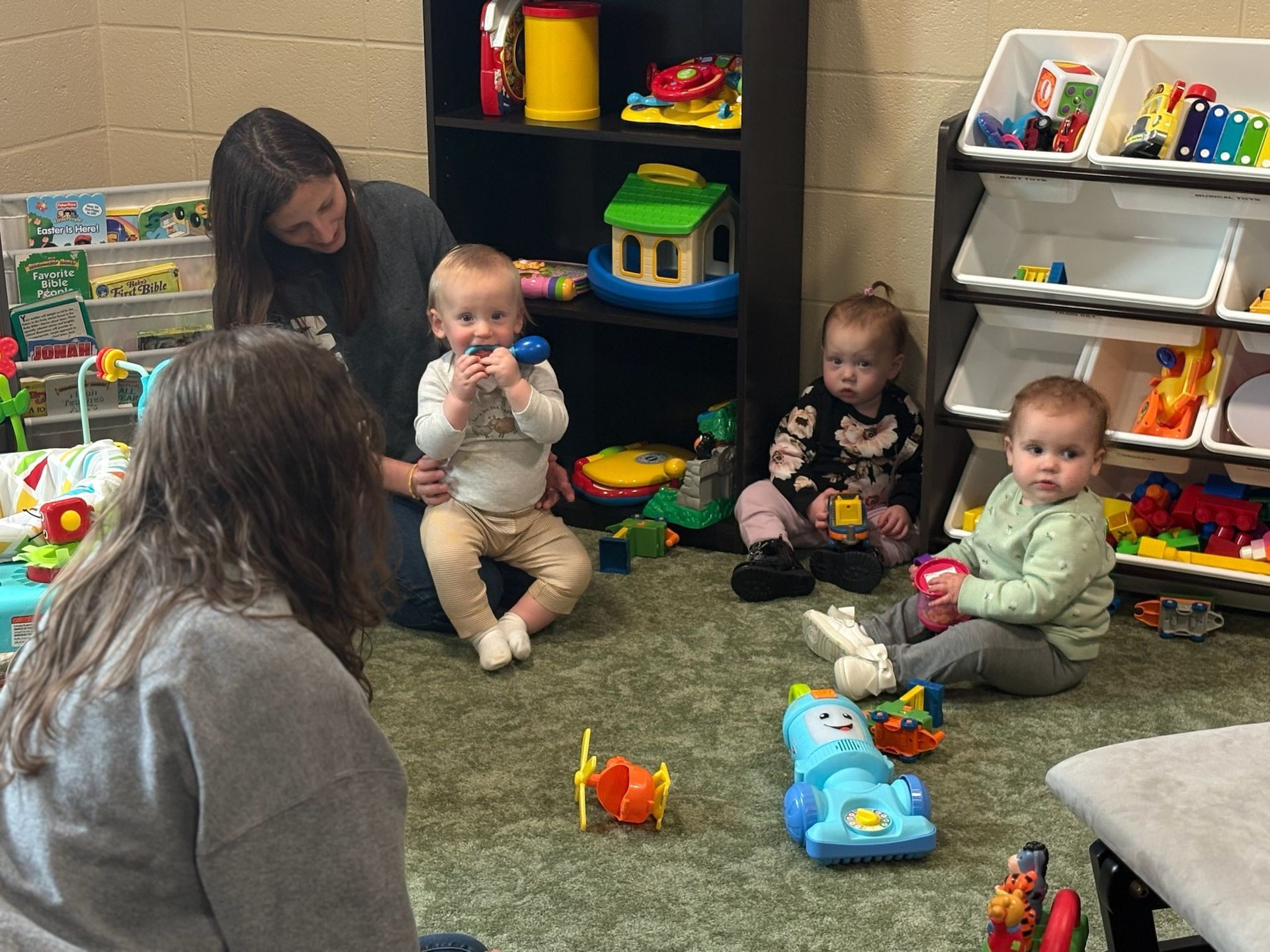 Adult and four babies sitting on a carpet in a daycare play area with toys and shelves nearby
