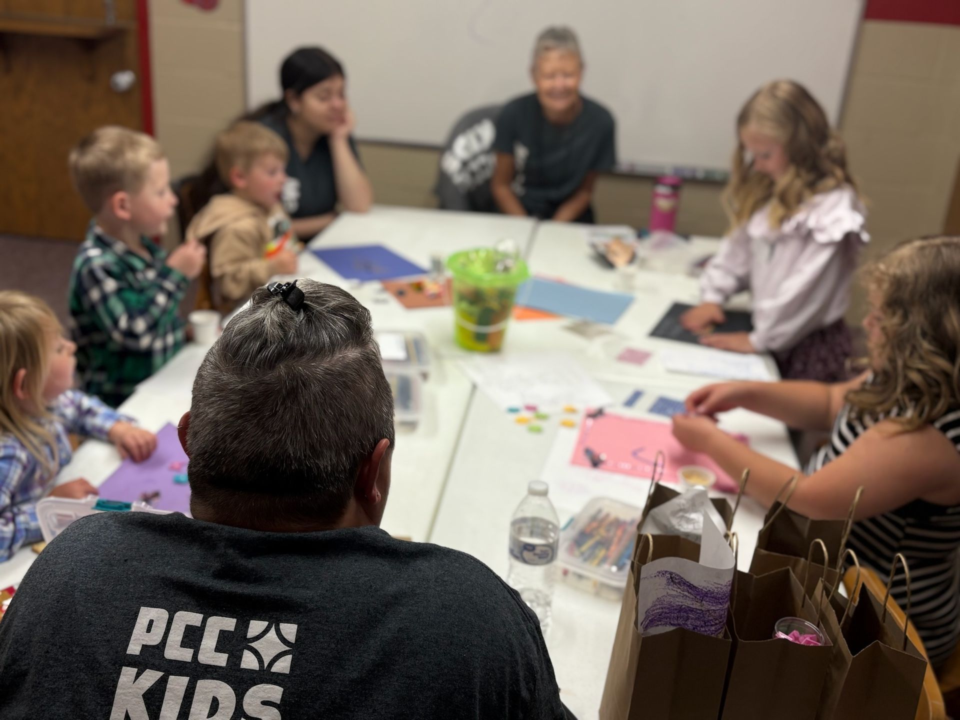 Children and adults seated around a table, drawing and crafting in a classroom or community activity room
