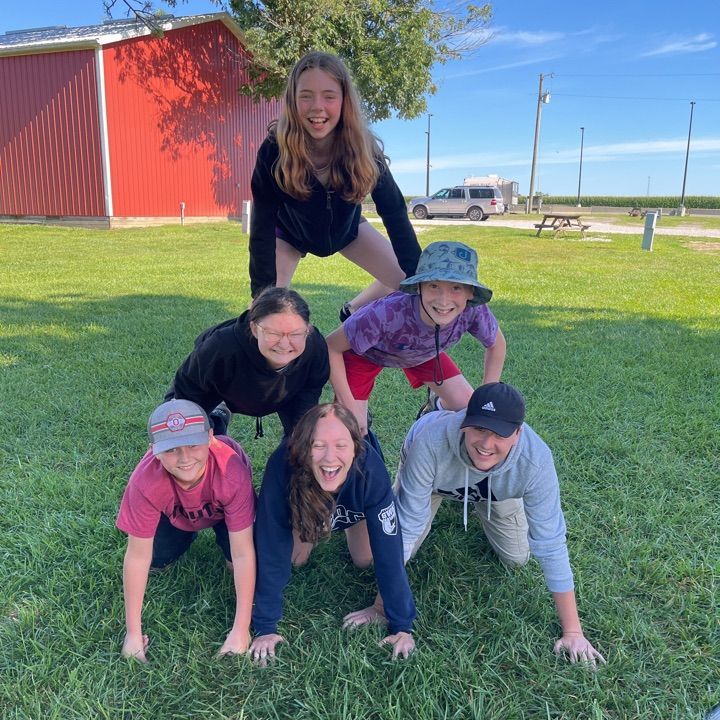 Six people pose in a human pyramid on a grassy lawn near a red barn under a sunny blue sky.