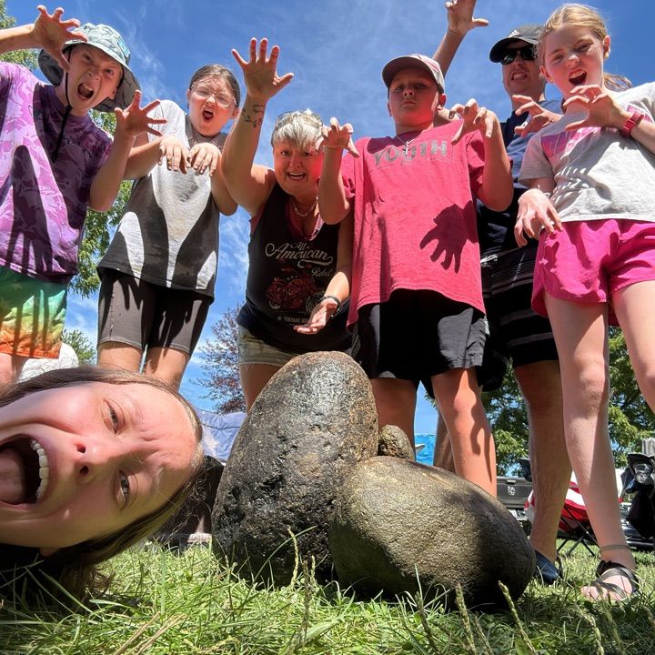 Children posing and smiling outdoors with raised hands around a pile of rocks on grass.