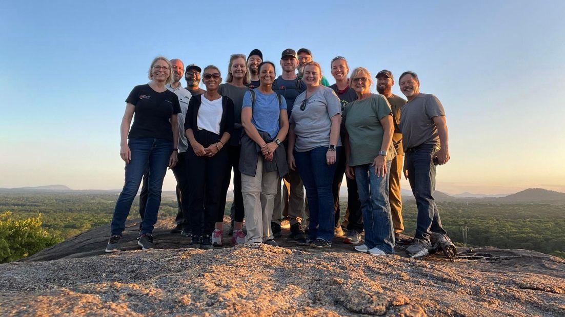 Group of people standing on a rocky hill at sunset, smiling with mountains in the background.