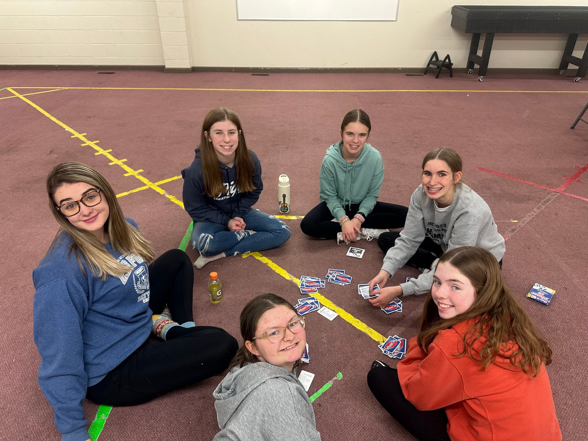 Five people sit on a gym floor around scattered game pieces, smiling during a board game.