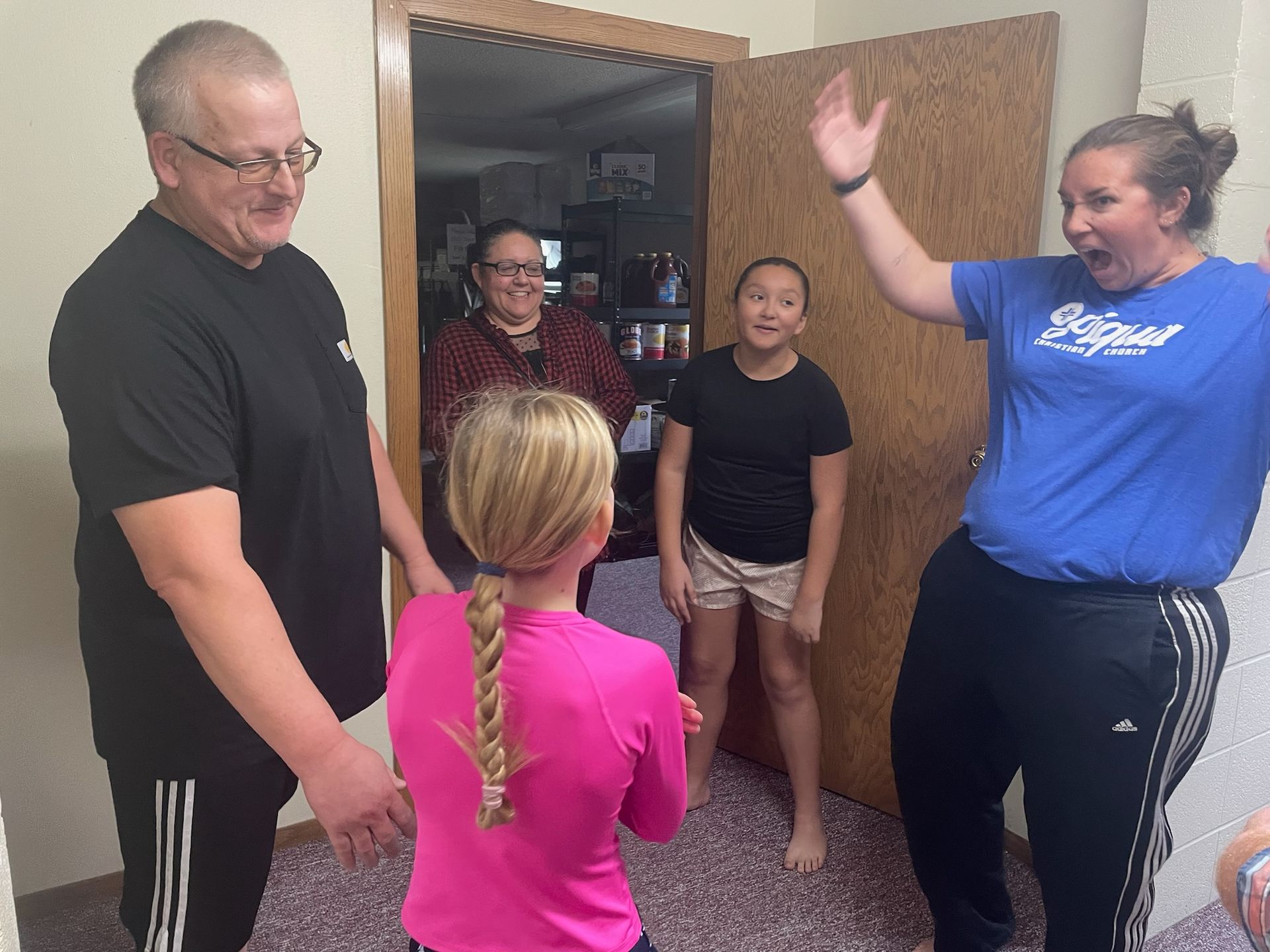 Five people standing in a doorway, smiling and gesturing as if greeting each other.