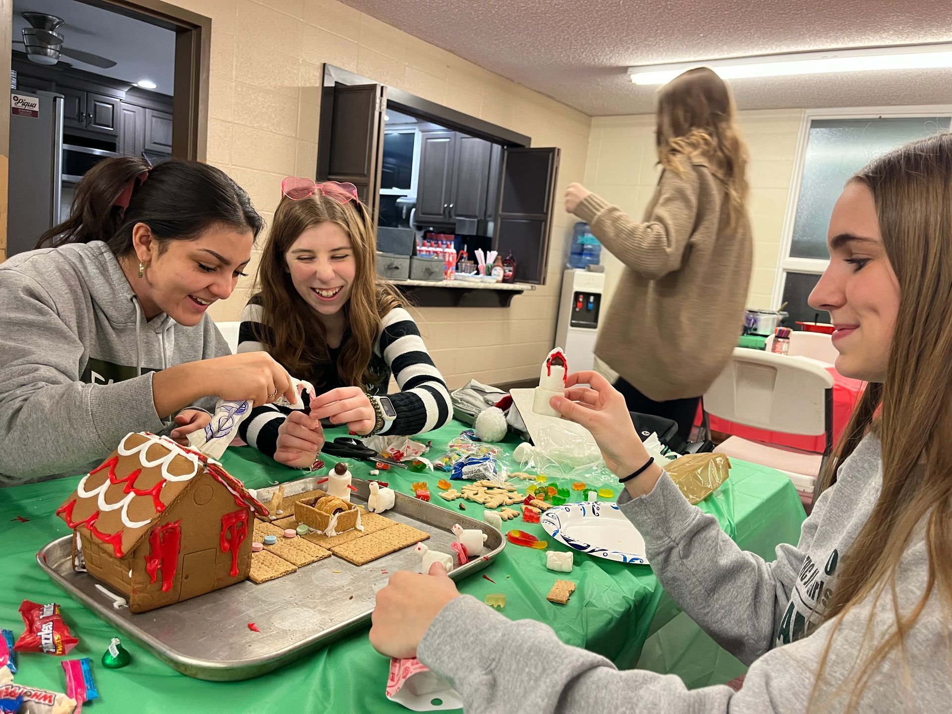 People decorating a gingerbread house at a festive table with candy and icing