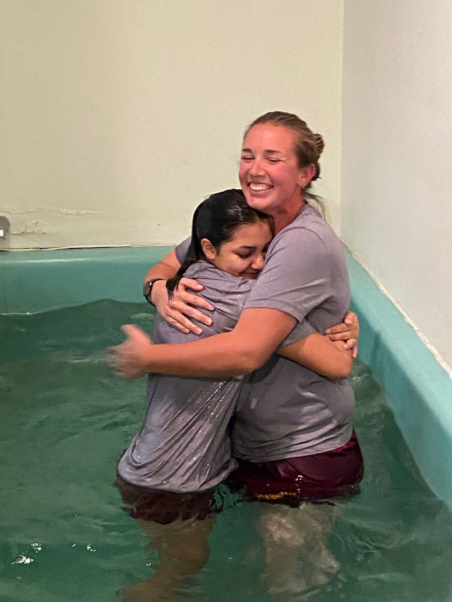 Two people hugging in a water tank, both smiling, with one wearing a gray shirt and maroon shorts.
