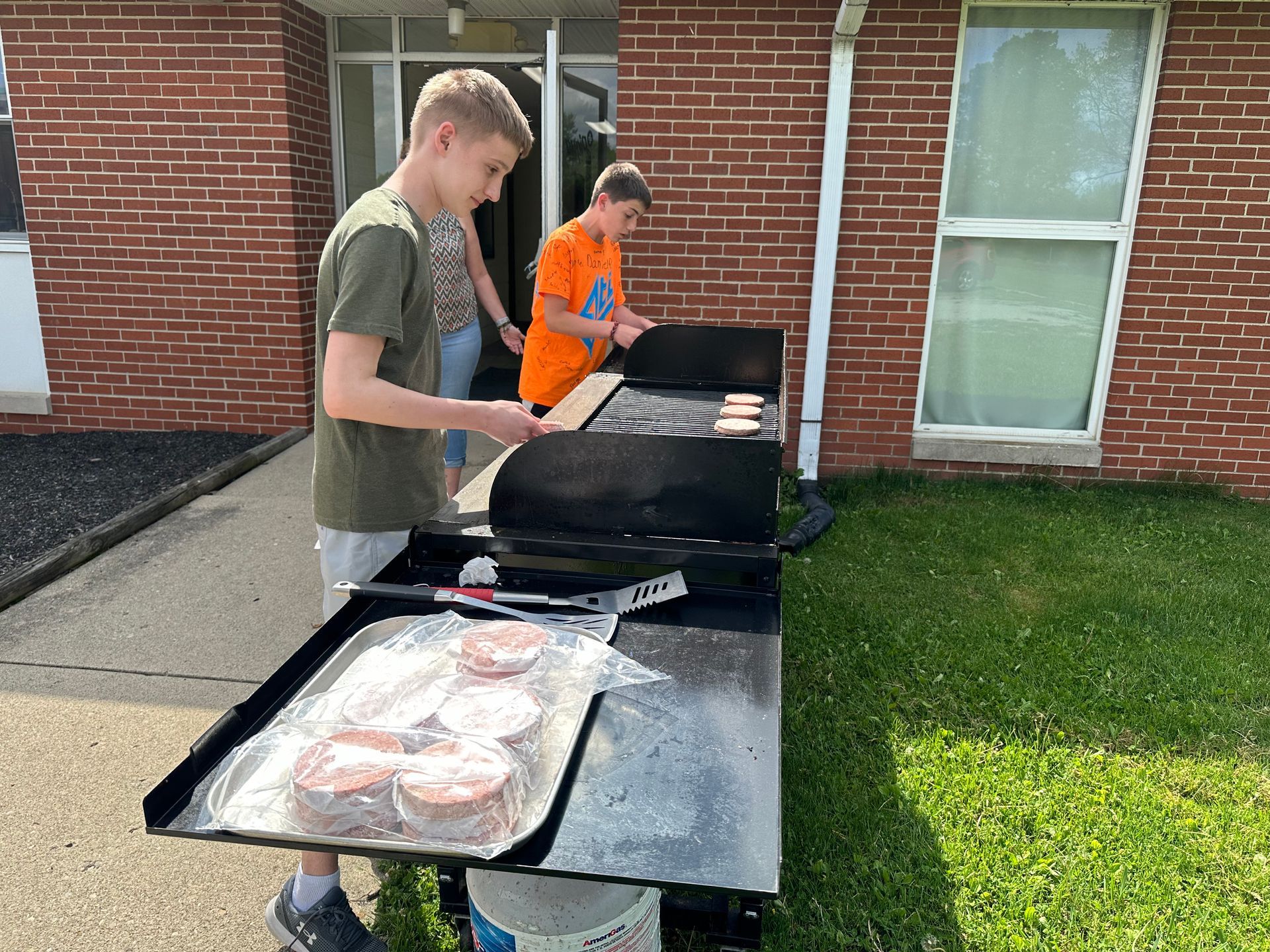 Two people grilling burgers at an outdoor picnic table beside a brick building