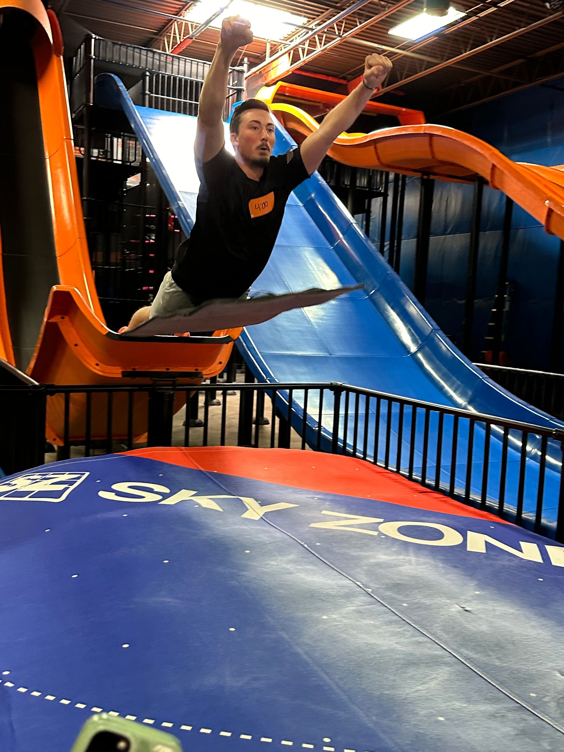 Person leaping off an orange slide in an indoor play area, landing on a blue mat.