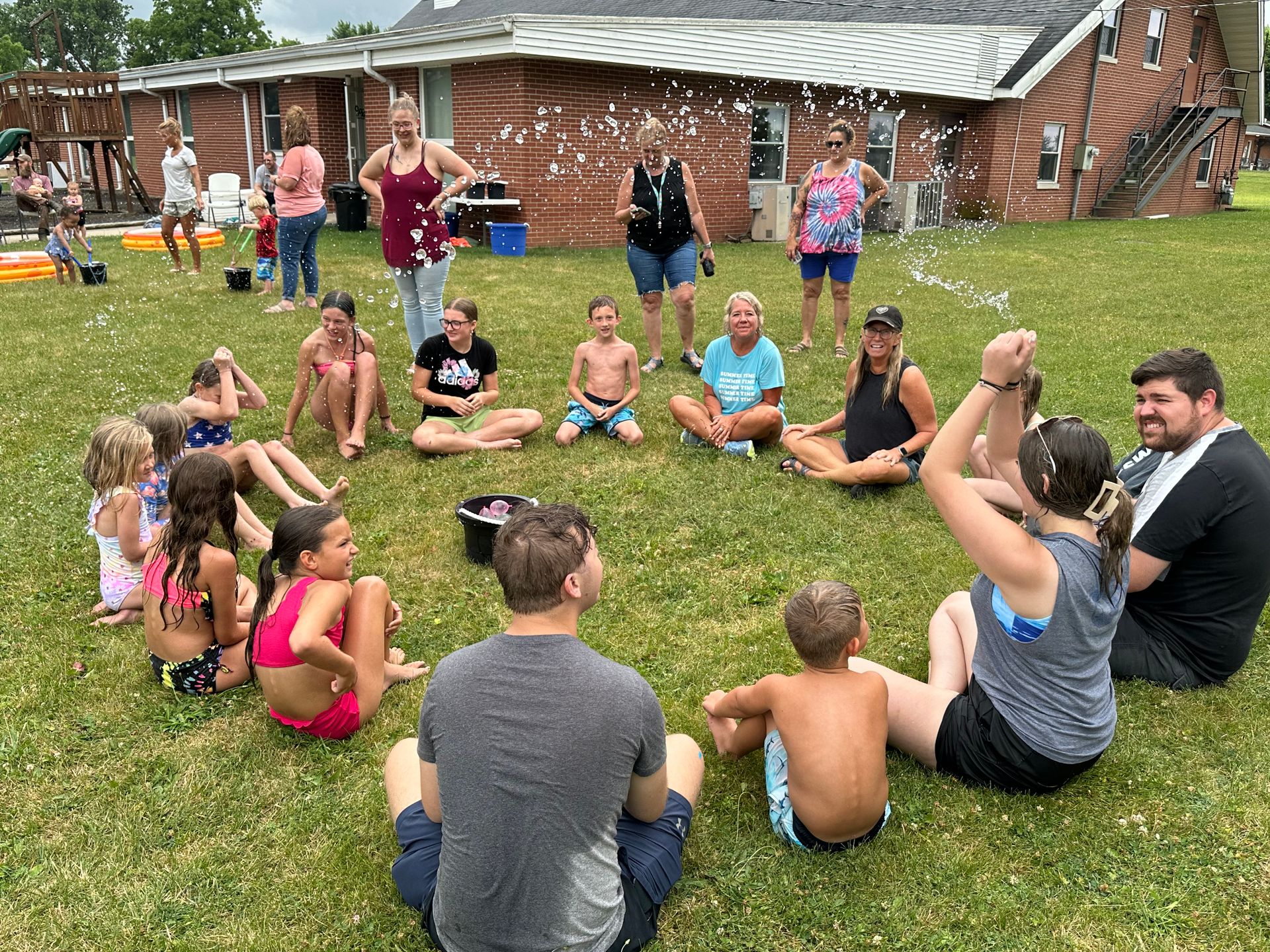 Group of people sitting in a circle on grass outdoors near cabins, listening during an activity