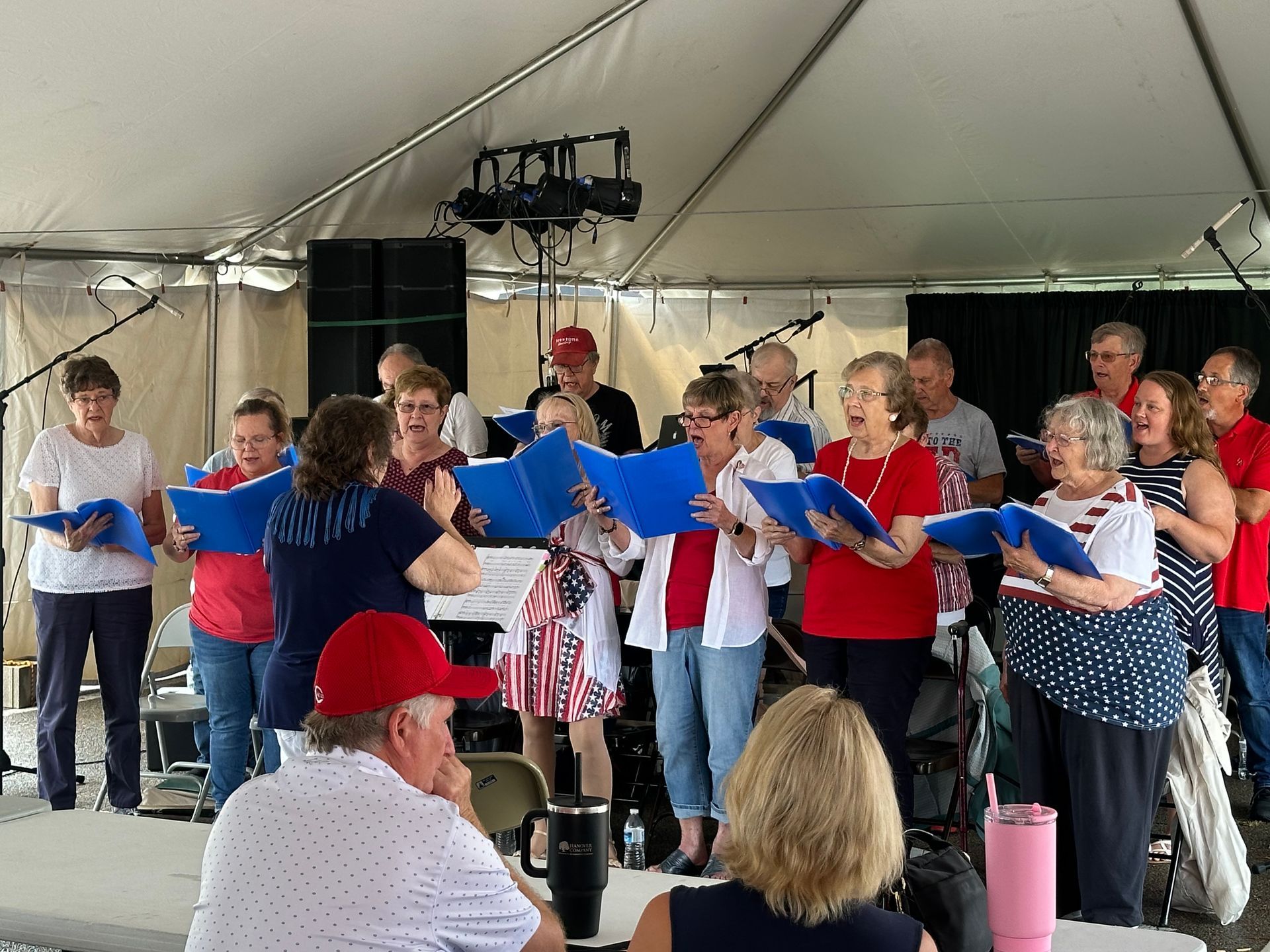Choir singing under a white tent, holding blue song sheets on a stage.
