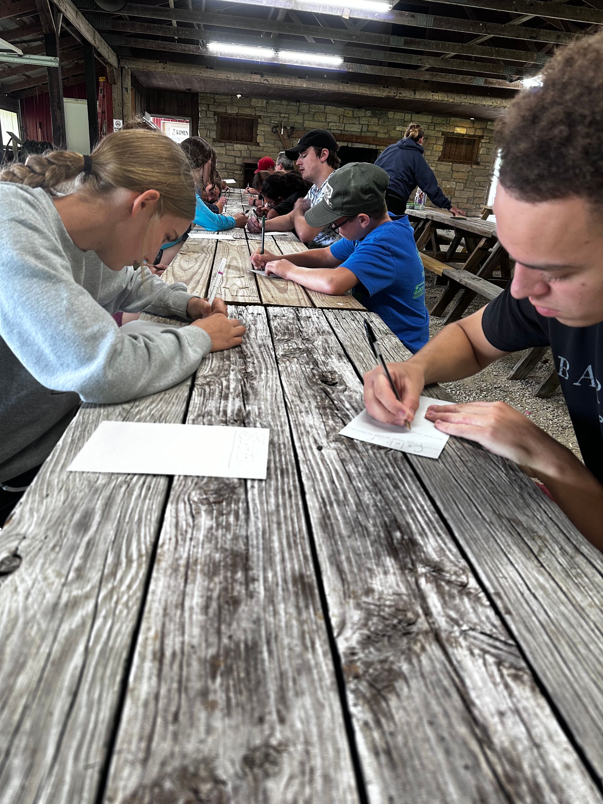 People seated at a long picnic table, writing on paper under a covered outdoor shelter.