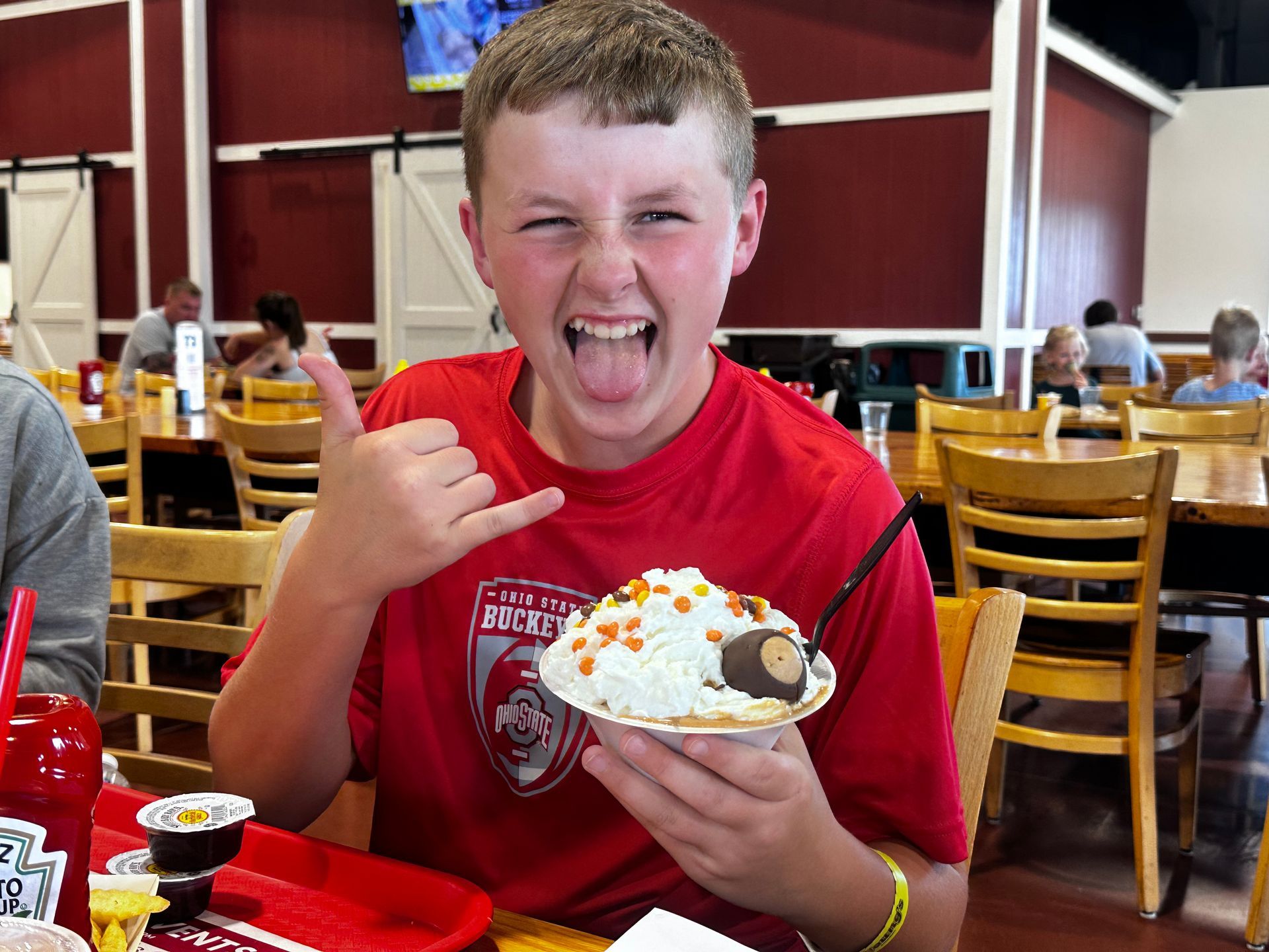 Boy in red shirt holding a sundae and making a playful shaka sign in a restaurant.