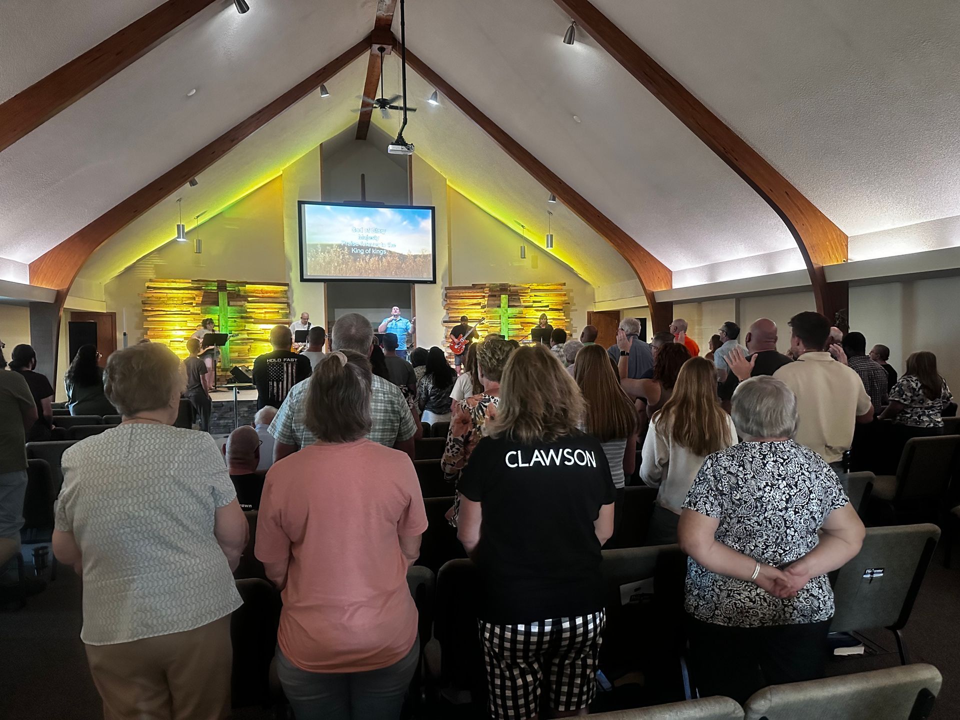 Congregation standing in a church hall, facing a stage with a projected screen and worship leaders.