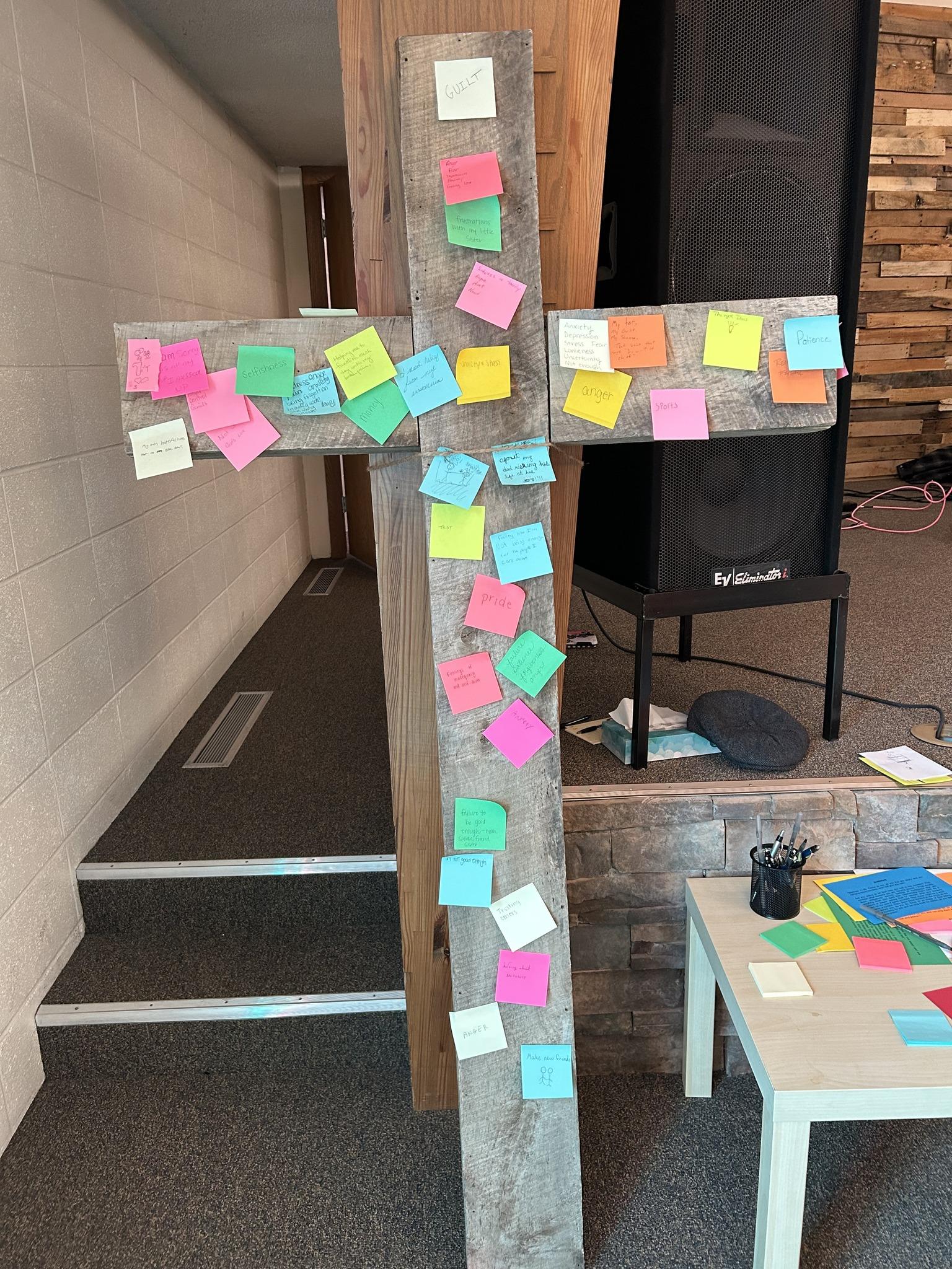 Wooden cross covered with colorful sticky notes in a hallway, with a small table nearby.