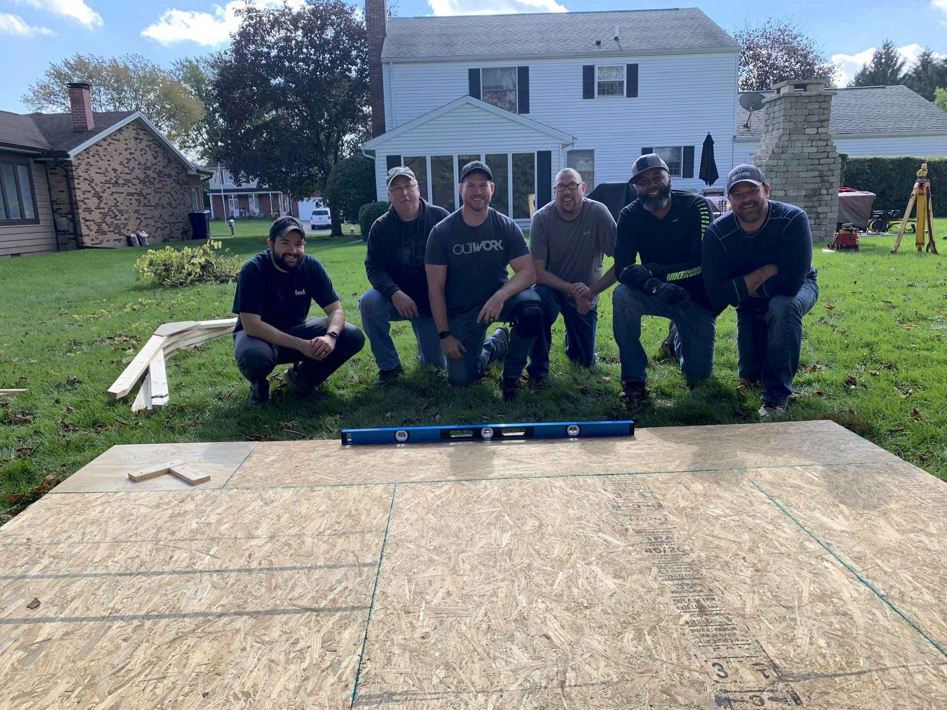 Five people kneel behind a wooden platform frame on a grassy yard with a house in the background.