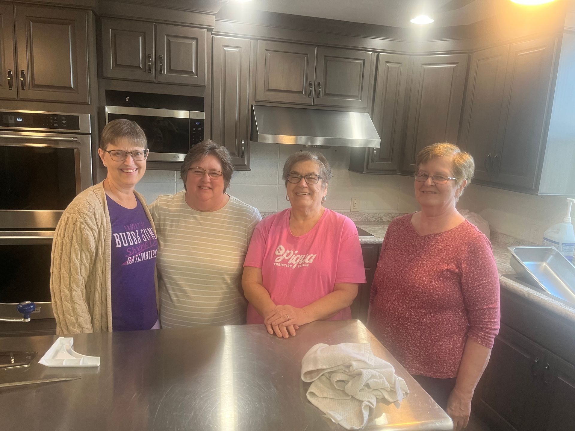 Four people stand in a kitchen behind a stainless counter, smiling at the camera.