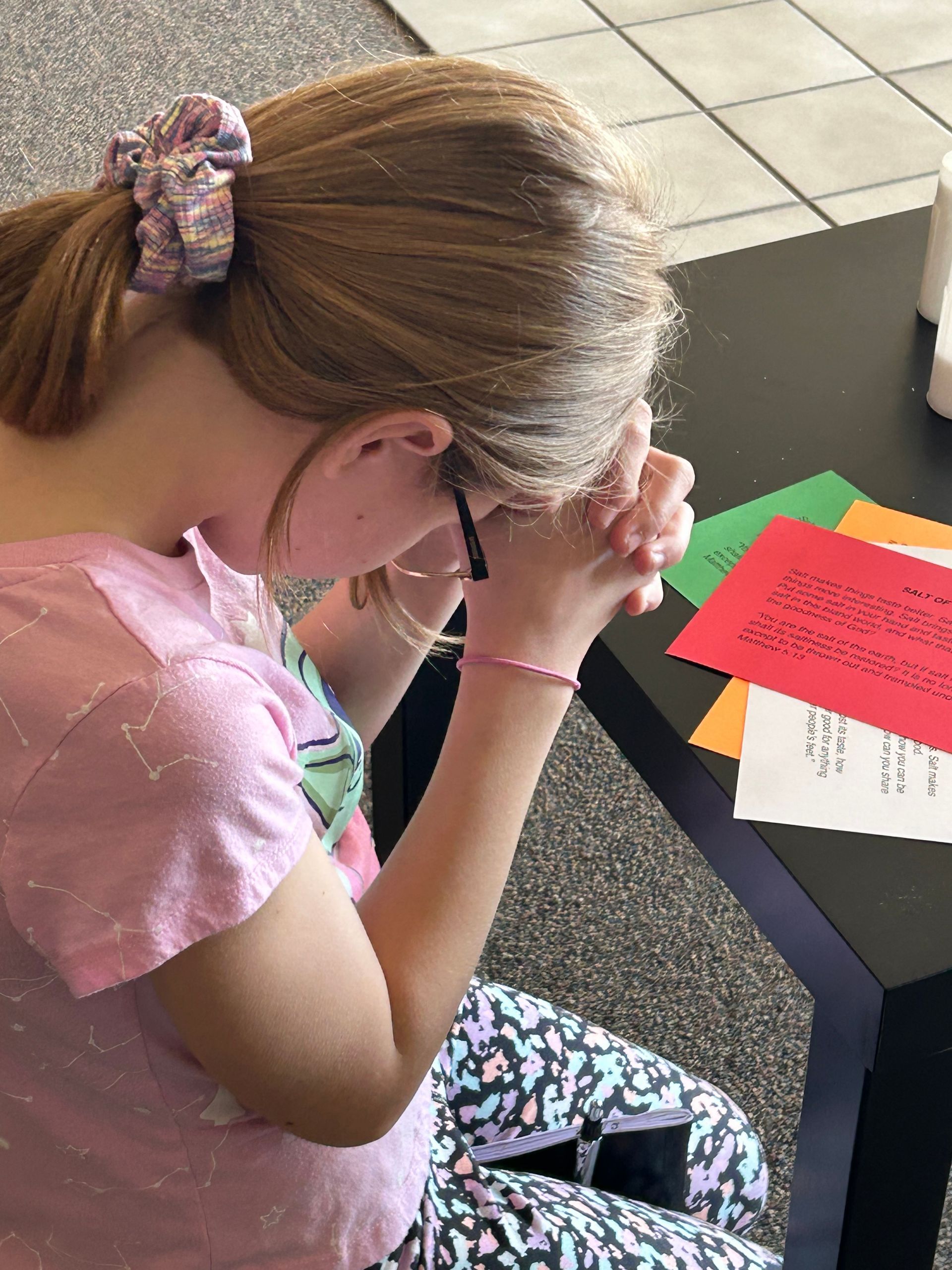 Child writing at a classroom desk with colorful papers and a pink shirt