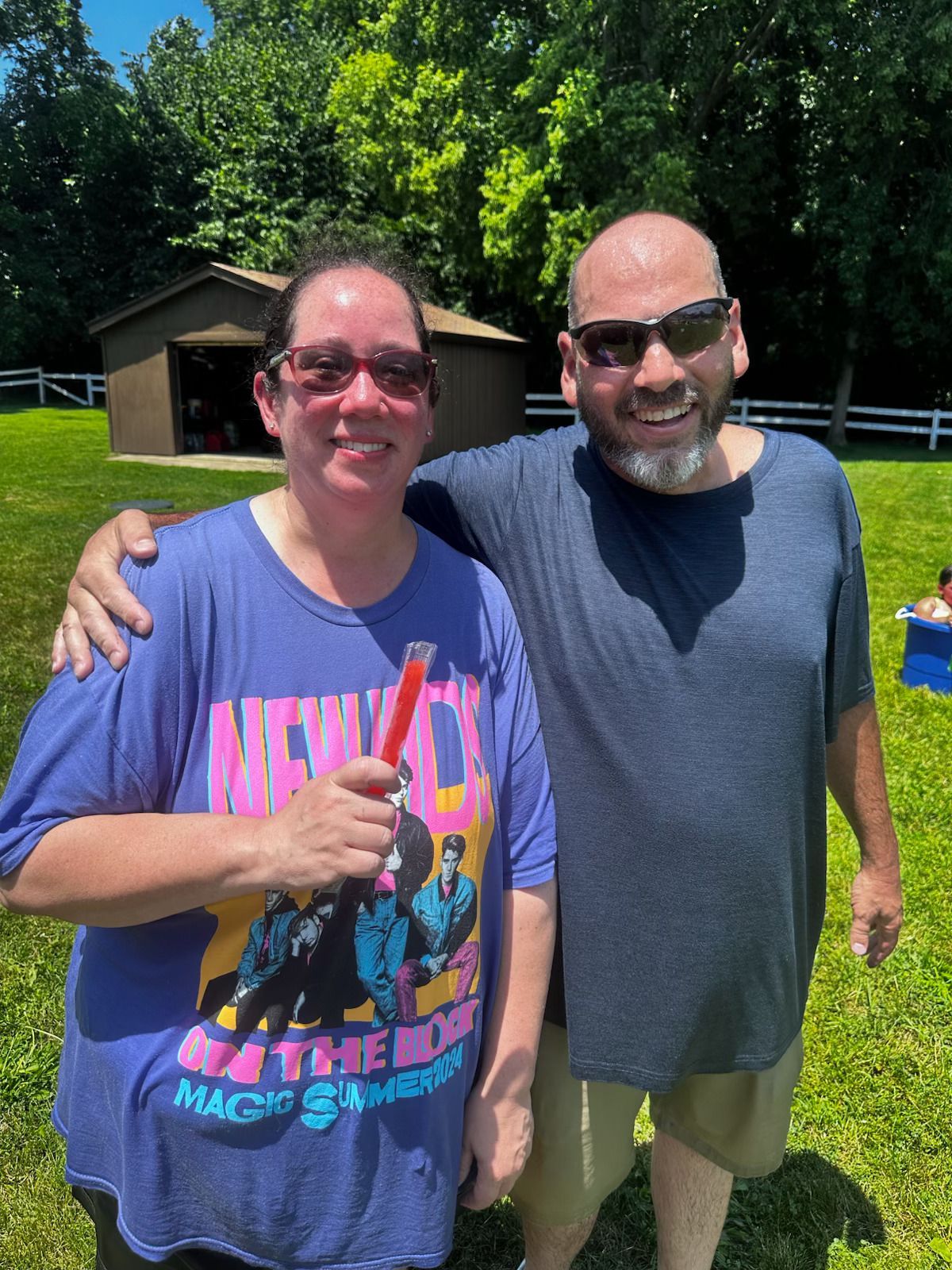 Two smiling people standing outdoors, one holding a thumbs-up, with trees and a small building behind them.