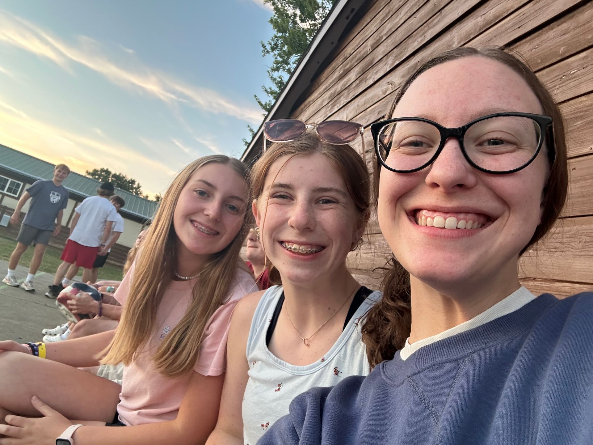 Three smiling friends sitting on a bench outdoors at sunset near a wooden building.