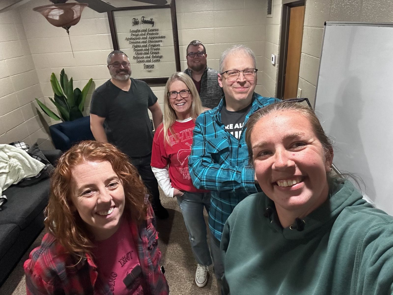 Group of five smiling people posing indoors, with a whiteboard and potted plant in the background.