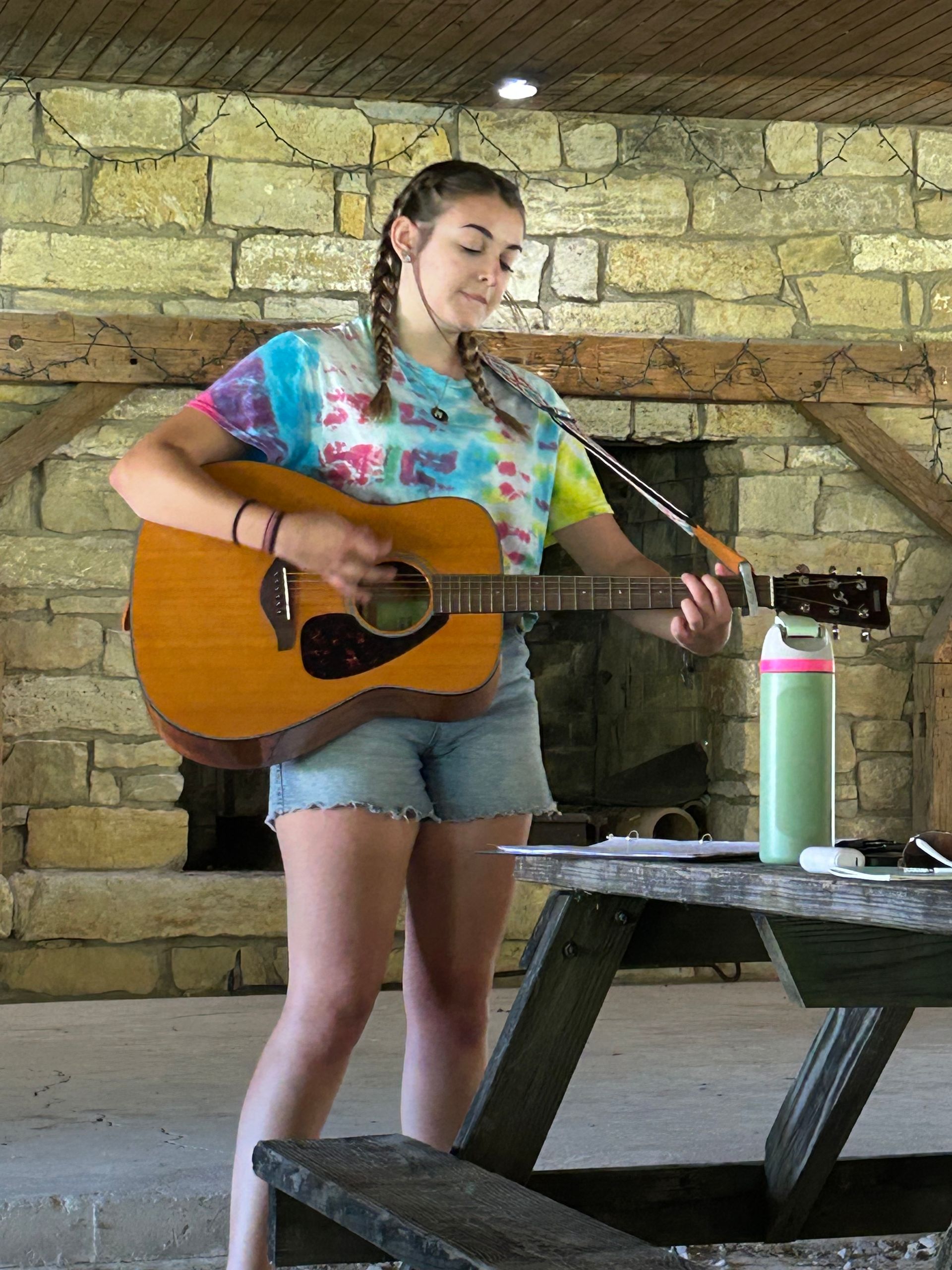 Person playing acoustic guitar in a stone pavilion beside a picnic table.