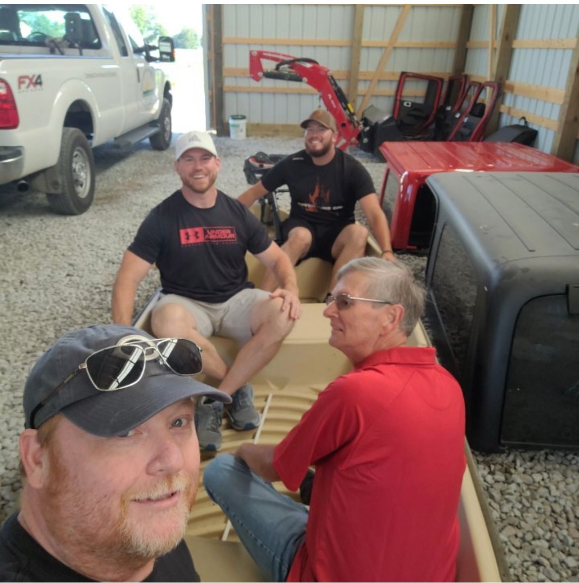 Four people sit and chat in a garage beside a red tractor and white pickup truck.