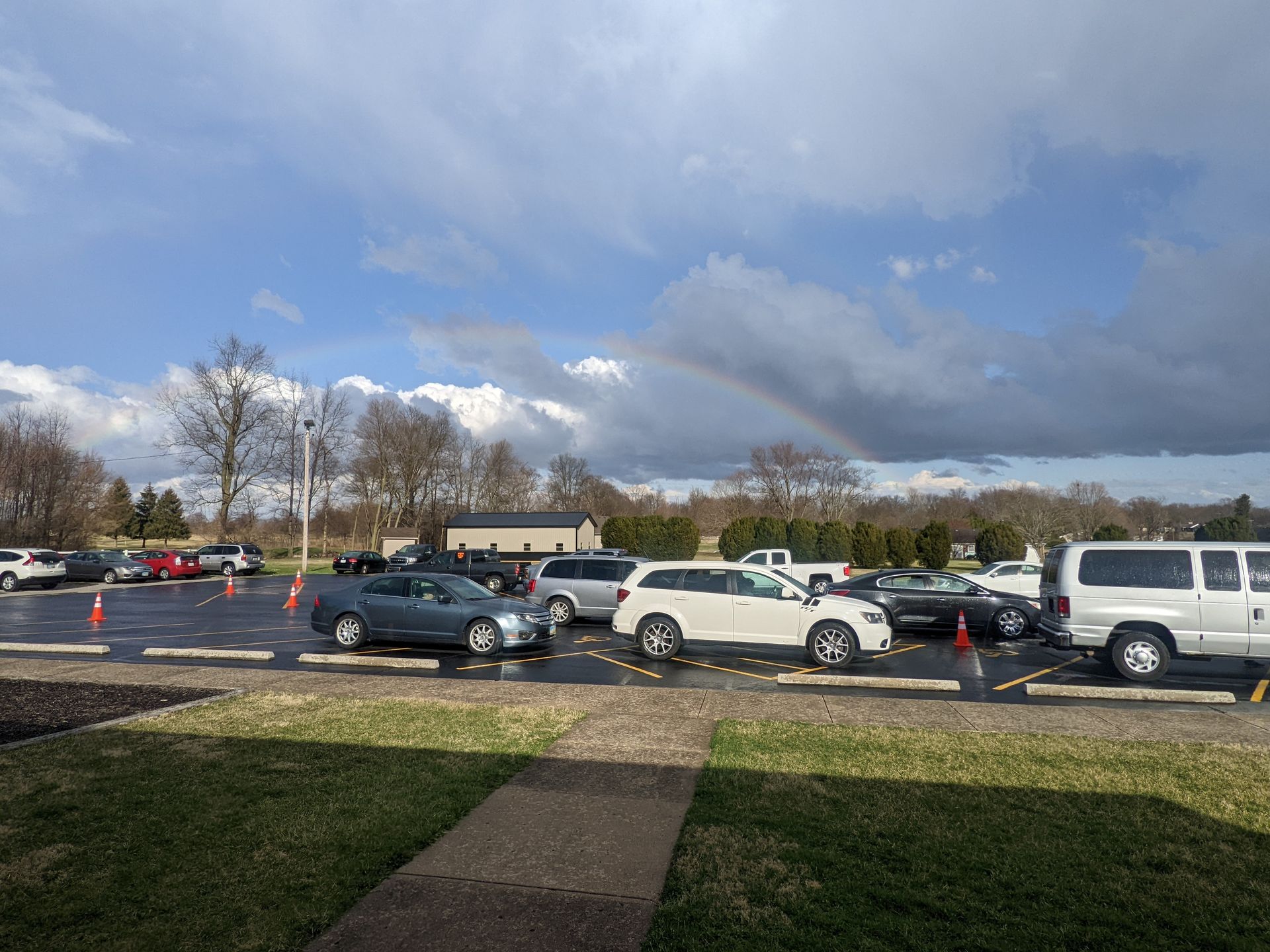Parking lot with several cars under a cloudy sky, viewed from a sidewalk and grassy area.