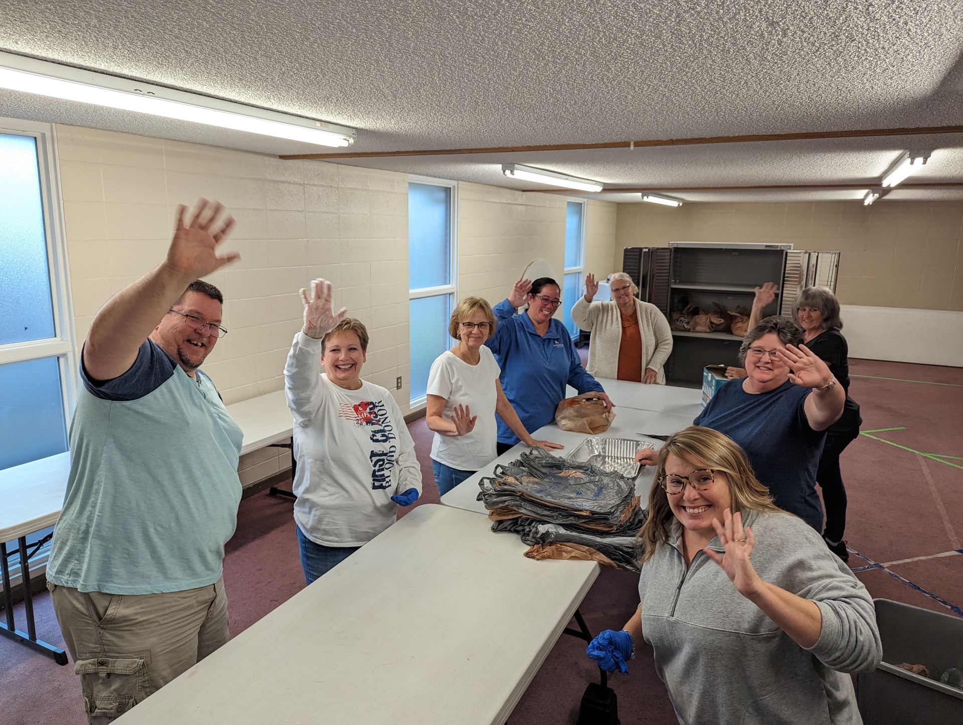 People waving around a long table in a bright room with fluorescent lights