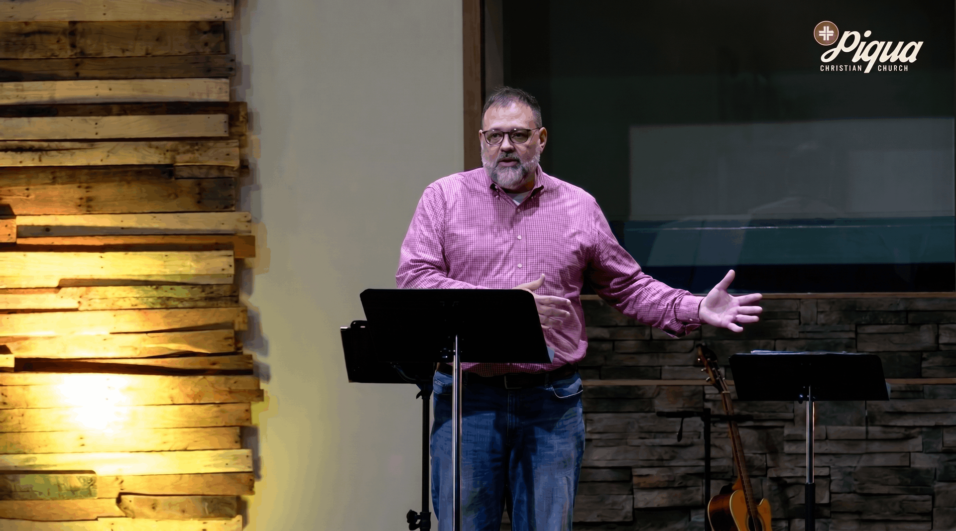 Man speaking at a podium on a stage, wearing a pink shirt with one arm extended, against a wood and stone backdrop.