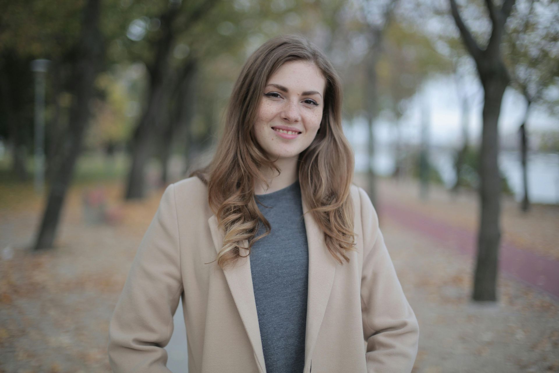 Woman in a beige coat smiling on a tree-lined path in autumn