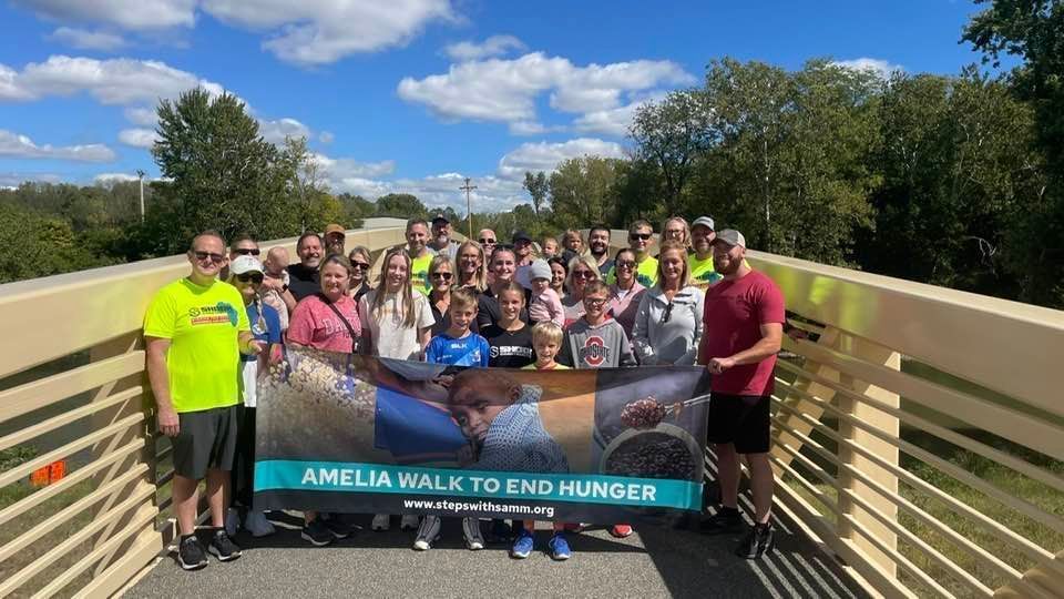 Group of walkers on a bridge holding a “Amelia Walk to End Hunger” banner under a sunny sky