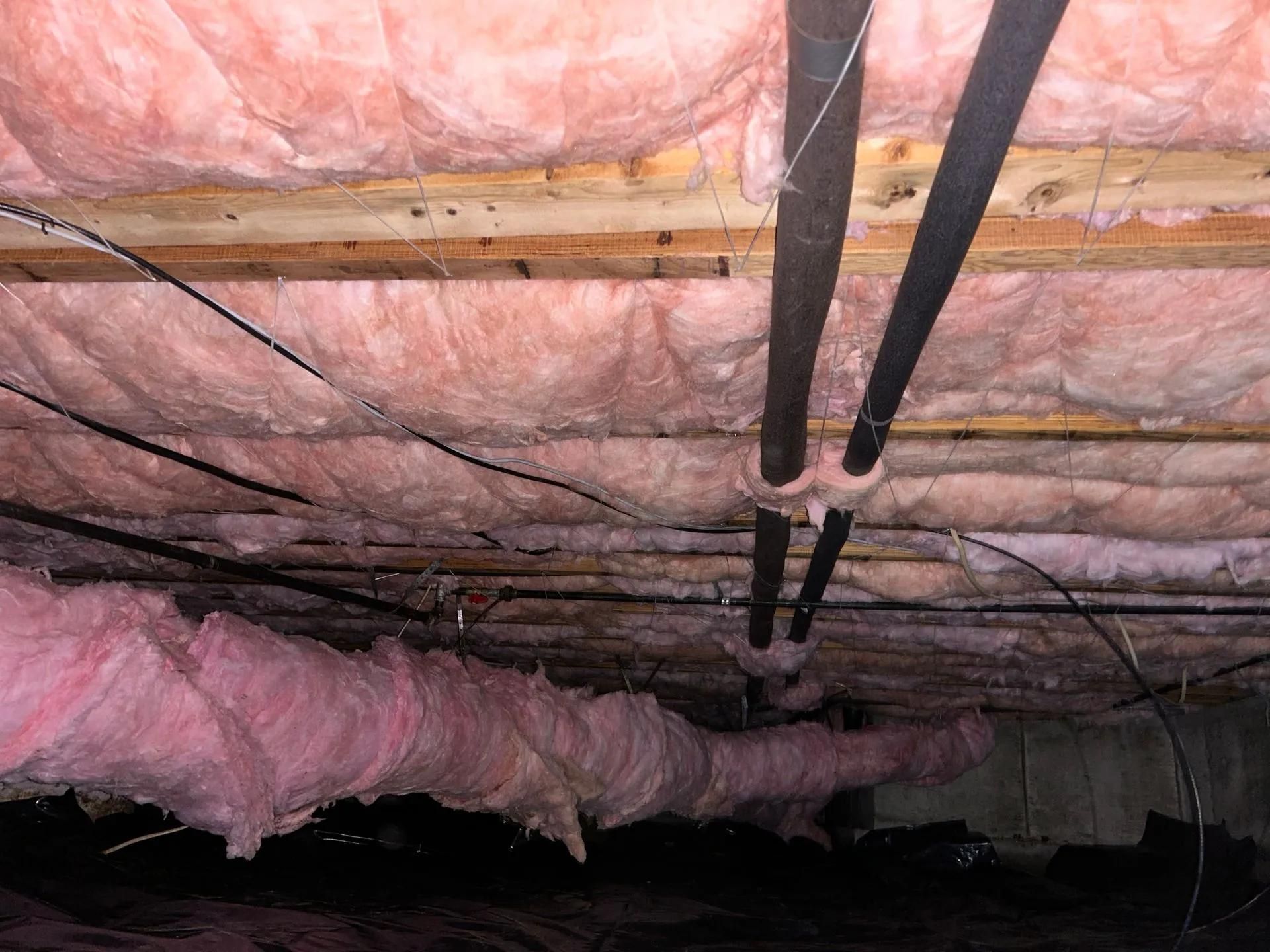 Exposed pink insulation and black pipes running across an unfinished basement ceiling.