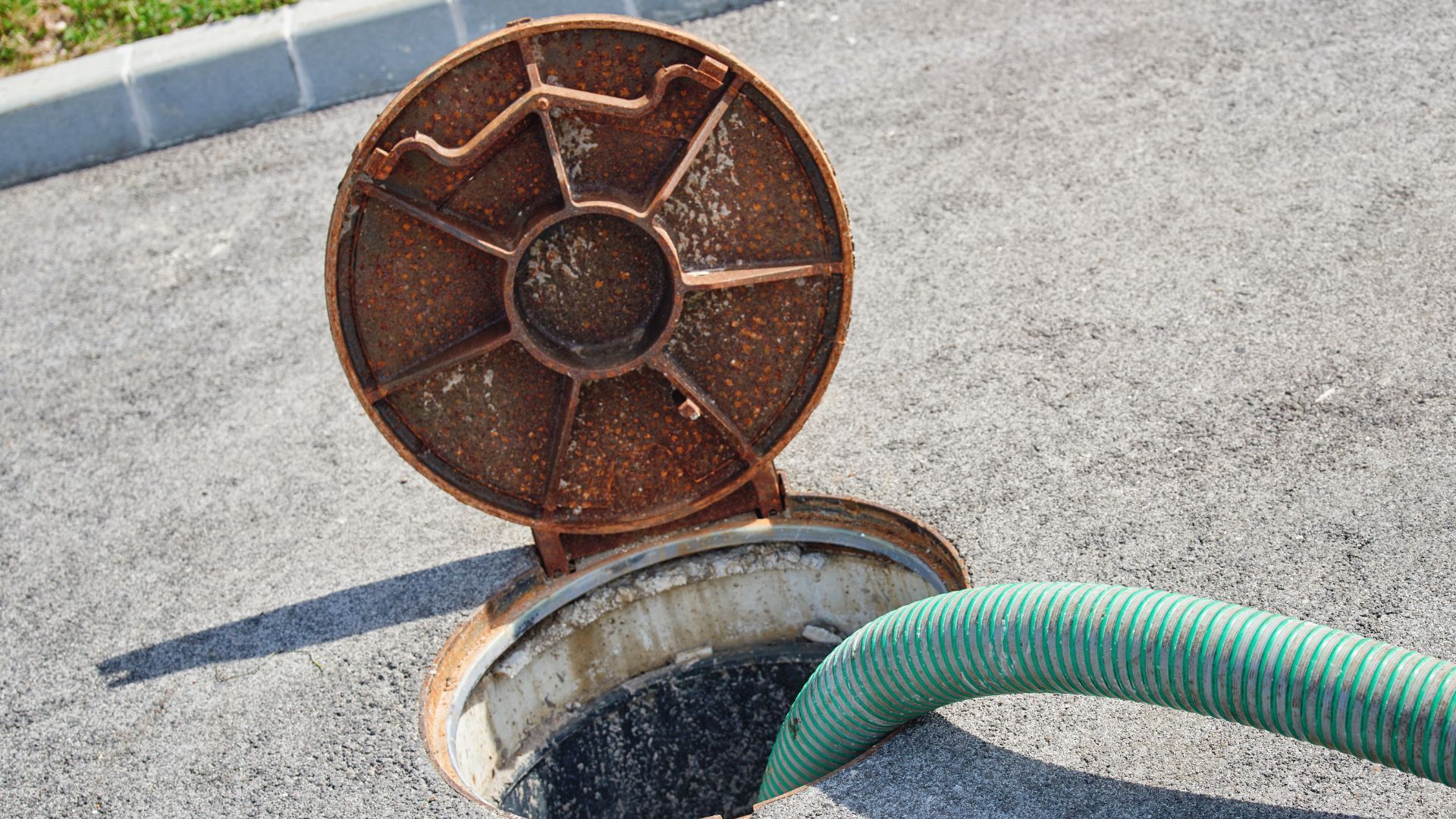 Open manhole with rusted circular lid and green hose on pavement
